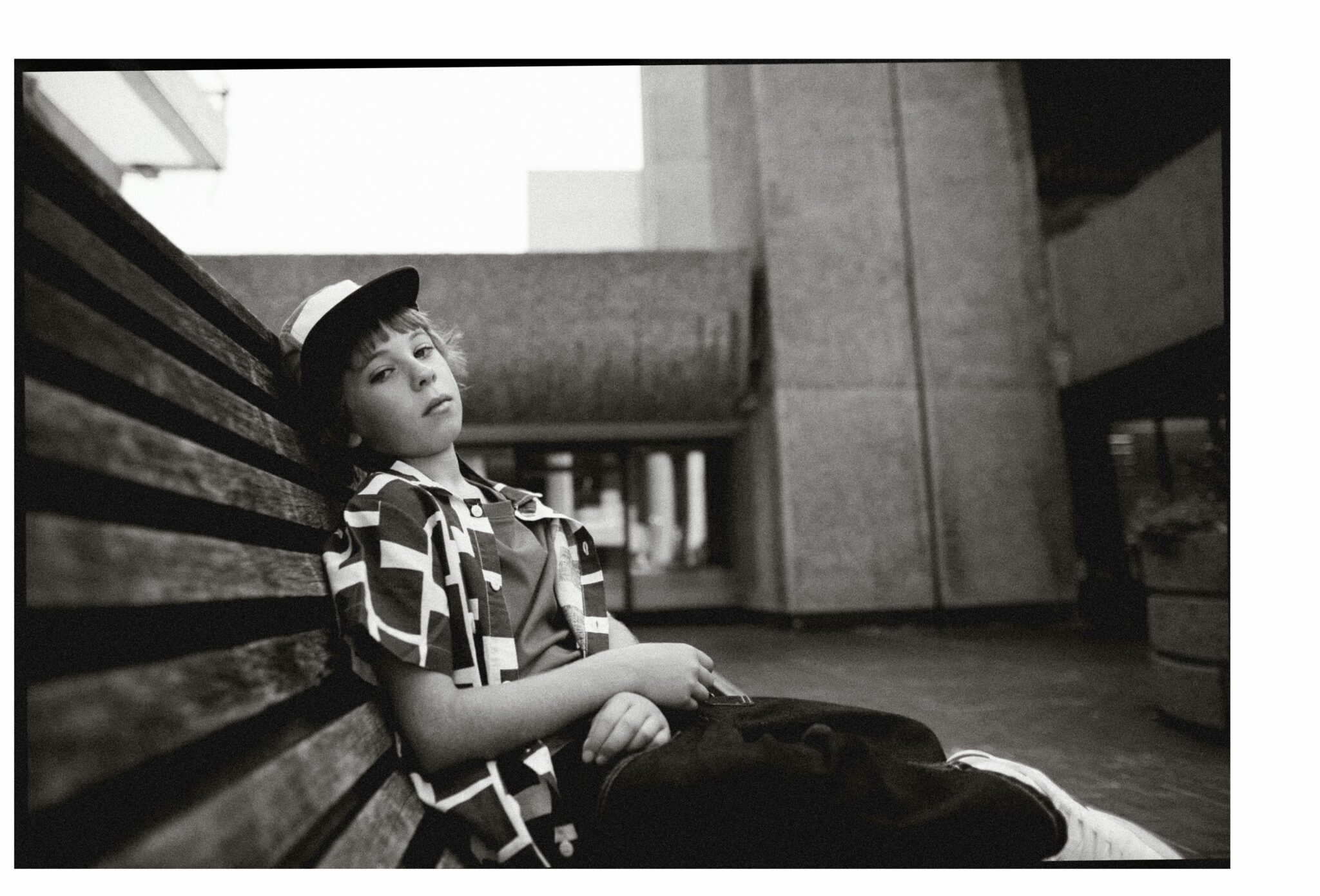 Sebastian leaning back on a wooden bench in an urban setting, wearing SS25 dark jeans by The New Society, graphic short-sleeve shirt by Wynken, red T-shirt by H&M, cap by Zara, and white trainers. Photo by Hannah Mentz for Scimparello Magazine.