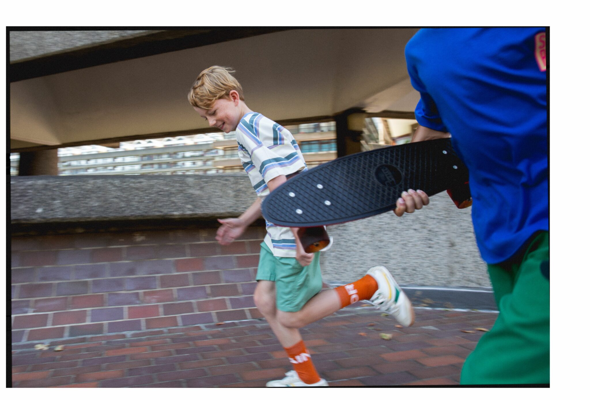 Sebastian running in an urban setting wearing SS25 look — striped oversized T-shirt by The Animals Observatory, green shorts by The New Society, rust orange socks by The Animals Observatory, and retro sneakers by Gucci, followed by Oliver in cobalt blue T-shirt and green trousers by The Animals Observatory holding a black skateboard. Photo by Hannah Mentz for Scimparello Magazine.