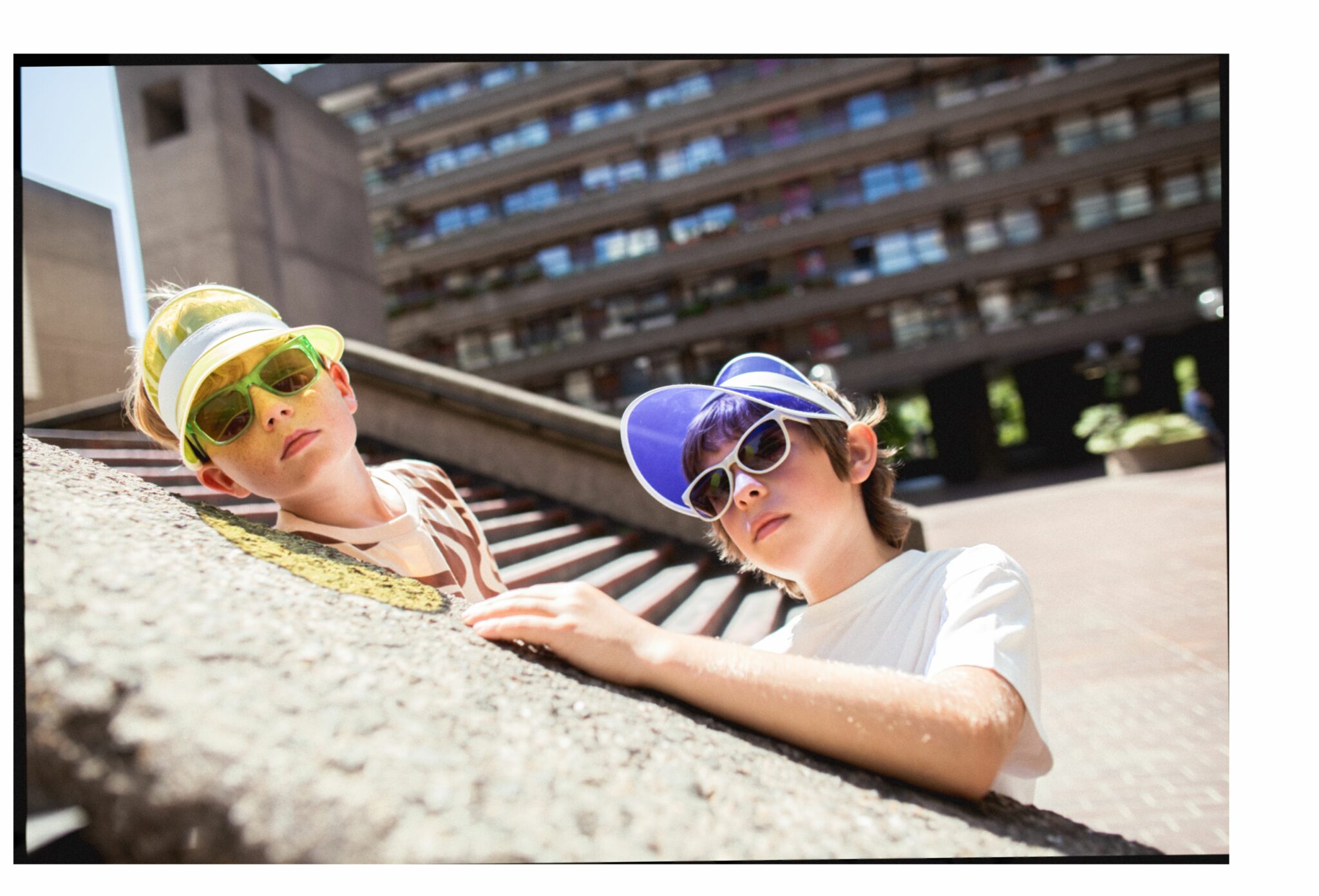Gene wearing SS25 brown and beige patterned T-shirt and neon green visor with matching sunglasses, next to Sebastian wearing SS25 white T-shirt, blue visor, and white retro sunglasses. They are leaning on a concrete slope in a sunny brutalist urban setting. Photo by Hannah Mentz for Scimparello Magazine.