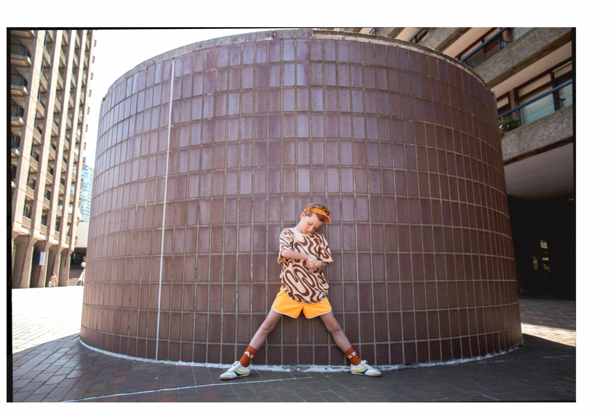 Gene posing with legs wide apart against a large curved wall covered in brown tiles, wearing SS25 brown and beige patterned T-shirt and orange socks by MAIN STORY, bright orange shorts, visor, and retro trainers by ZARA KIDS. Urban outdoor location with geometric architecture. Photo by Hannah Mentz for Scimparello Magazine.