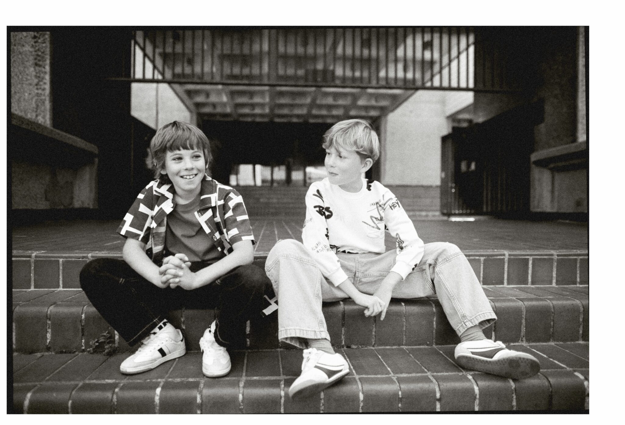 Sebastian wearing SS25 jeans by THE NEW SOCIETY, socks by MAIN STORY, short-sleeved shirt by WYNKEN, and T-shirt by H&M, sitting on brick steps next to Gene wearing SS25 jeans, sweatshirt, and trainers by ZARA, with socks by MAIN STORY. Outdoor urban scene. Photo by Hannah Mentz for Scimparello Magazine.