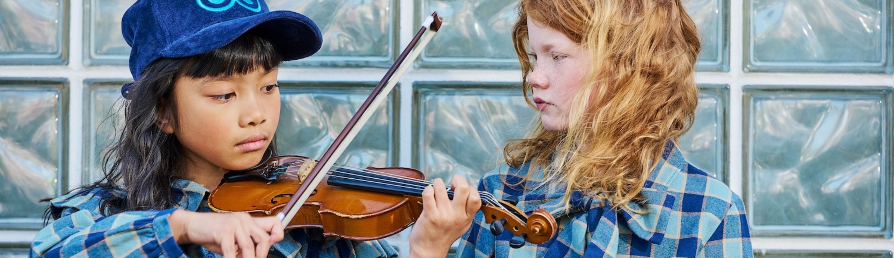 Two children in blue checkered outfits, one playing the violin and the other watching closely, against a glass block wall – LOUD FW25.