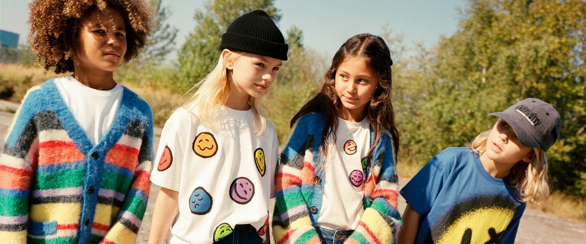 Four children outdoors wearing MOLO FW25 – colorful striped mohair cardigans, smiley print T-shirts, and bold graphic tops, styled with casual accessories.