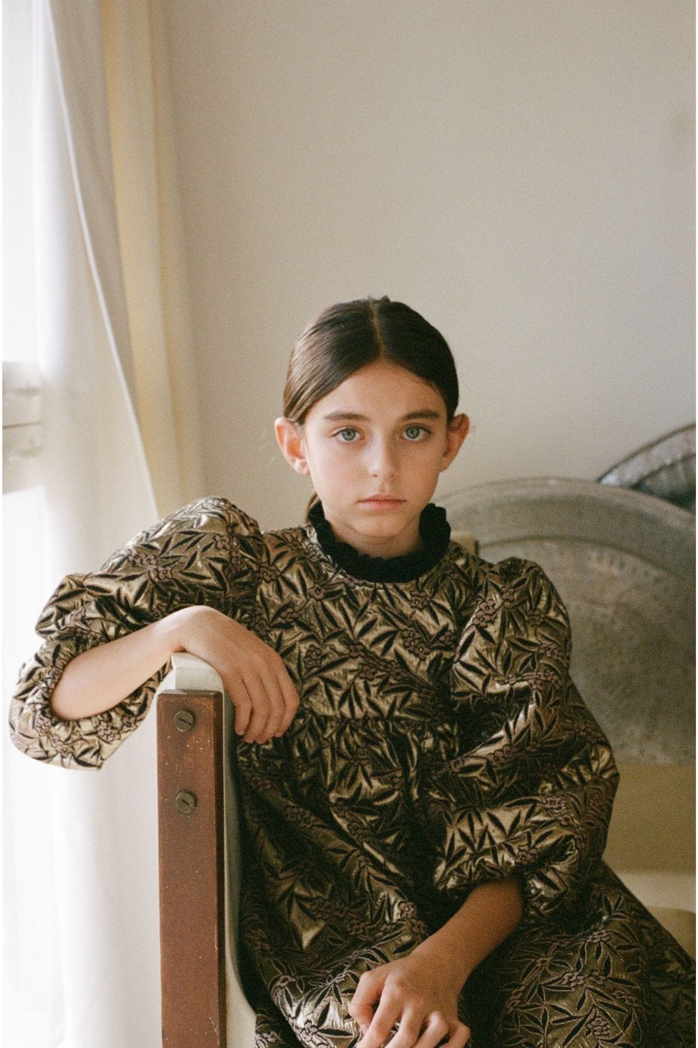Portrait of a girl sitting indoors wearing a metallic patterned dress with puff sleeves from Bebe Organic FW25. Photo featured on Scimparello Magazine.