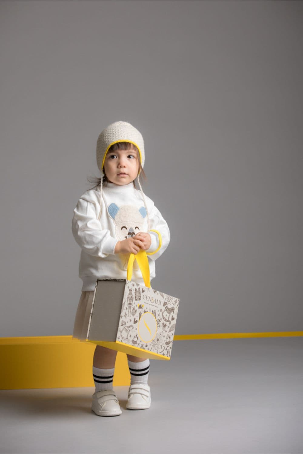 Toddler girl wearing cream knit bonnet and white bear sweater from Gensami FW25, holding a Gensami gift box with yellow handles.