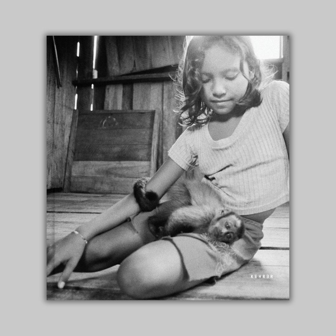 Cover of the book The Enchanted Ones by Stephanie Pommez, published by Kehrer Verlag and featured on Scimparello Magazine. Black and white portrait of a Ribeirinha girl sitting on a wooden floor with a capuchin monkey on her lap.