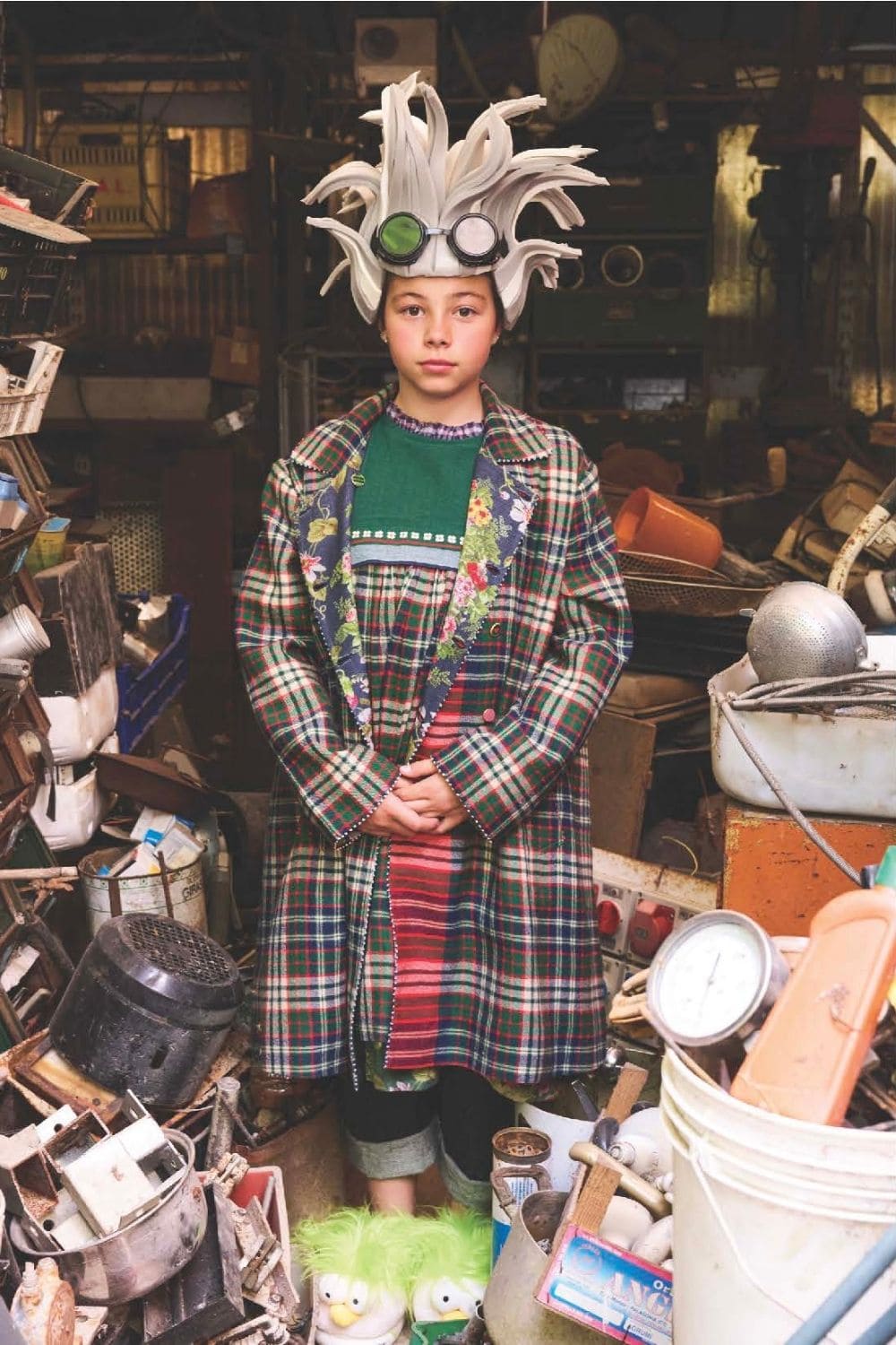Child in plaid coat with floral embroidery and green sweater, wearing playful white spiky wig with goggles, surrounded by vintage objects, PÉRO FW25 new collection on Scimparello Magazine.