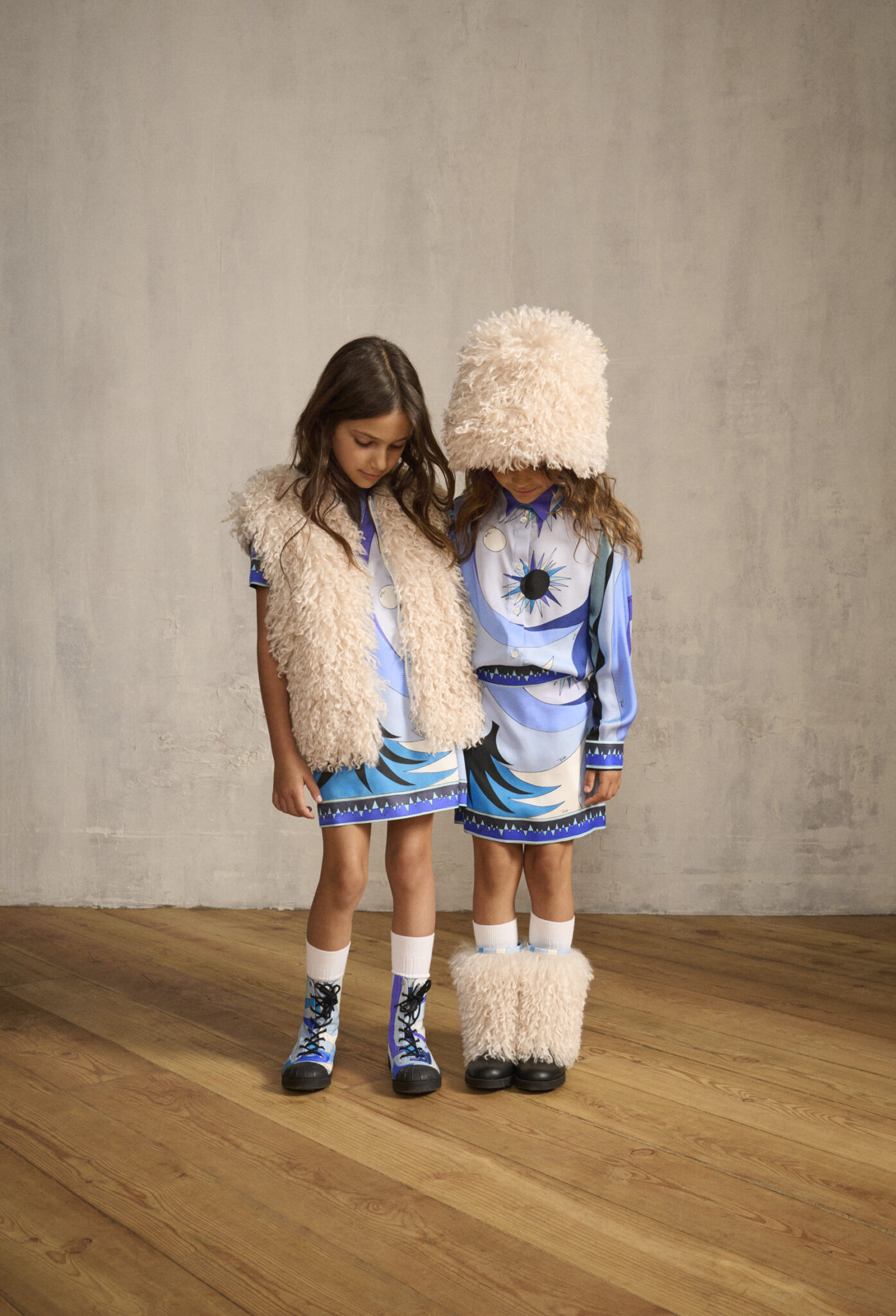 Two girls in FW25 Pucci Junior outfits with blue printed dresses, one styled with a fluffy cream vest and the other with a matching oversized furry hat and boots. Photo for Scimparello Magazine.
