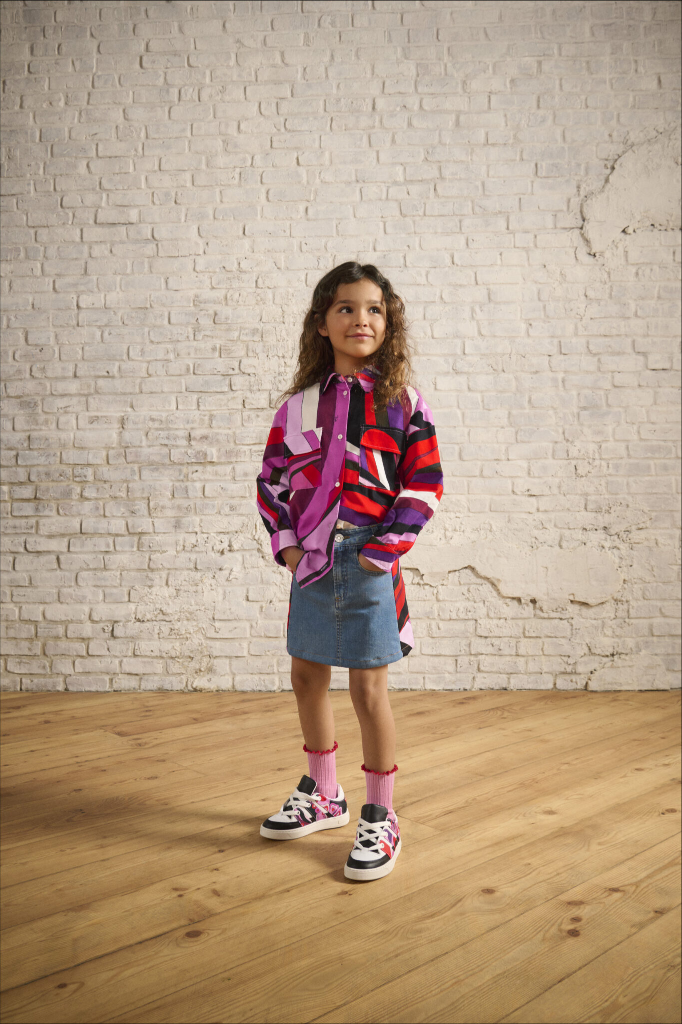Girl wearing FW25 Pucci Junior oversized striped shirt in red, purple, and pink, styled with a denim mini skirt and sneakers, standing against a white brick wall. Photo for Scimparello Magazine.