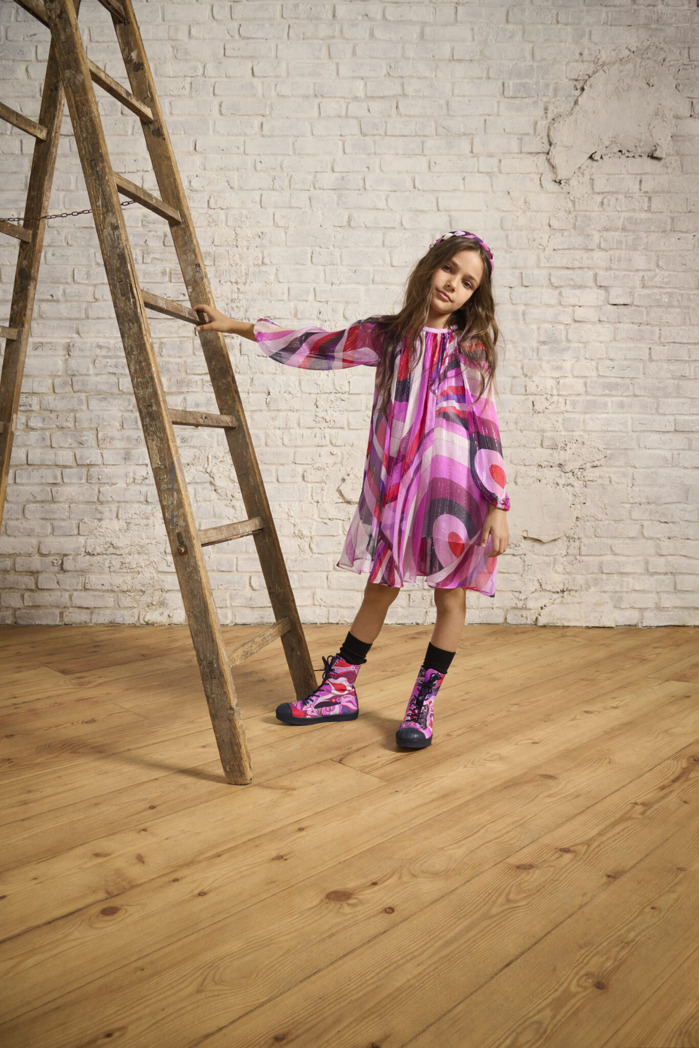 Girl in a flowing FW25 Pucci Junior dress with swirling pink and purple prints, styled with matching headband and printed boots, posing next to a rustic wooden ladder. Photo for Scimparello Magazine.