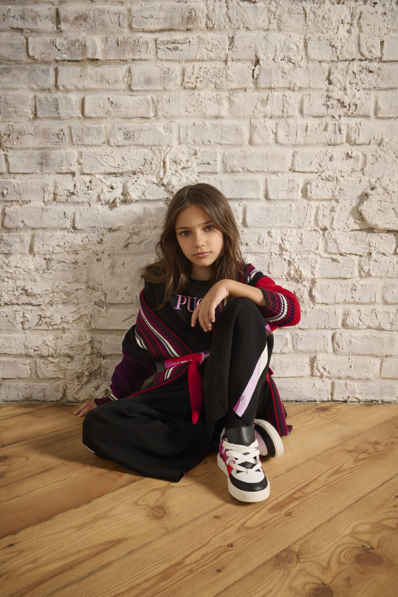 Girl sitting on the floor against a brick wall, wearing a FW25 Pucci Junior striped knit cardigan in red, black, and purple over a PUCCI T-shirt, styled with black trousers and sneakers, featured in Scimparello Magazine.