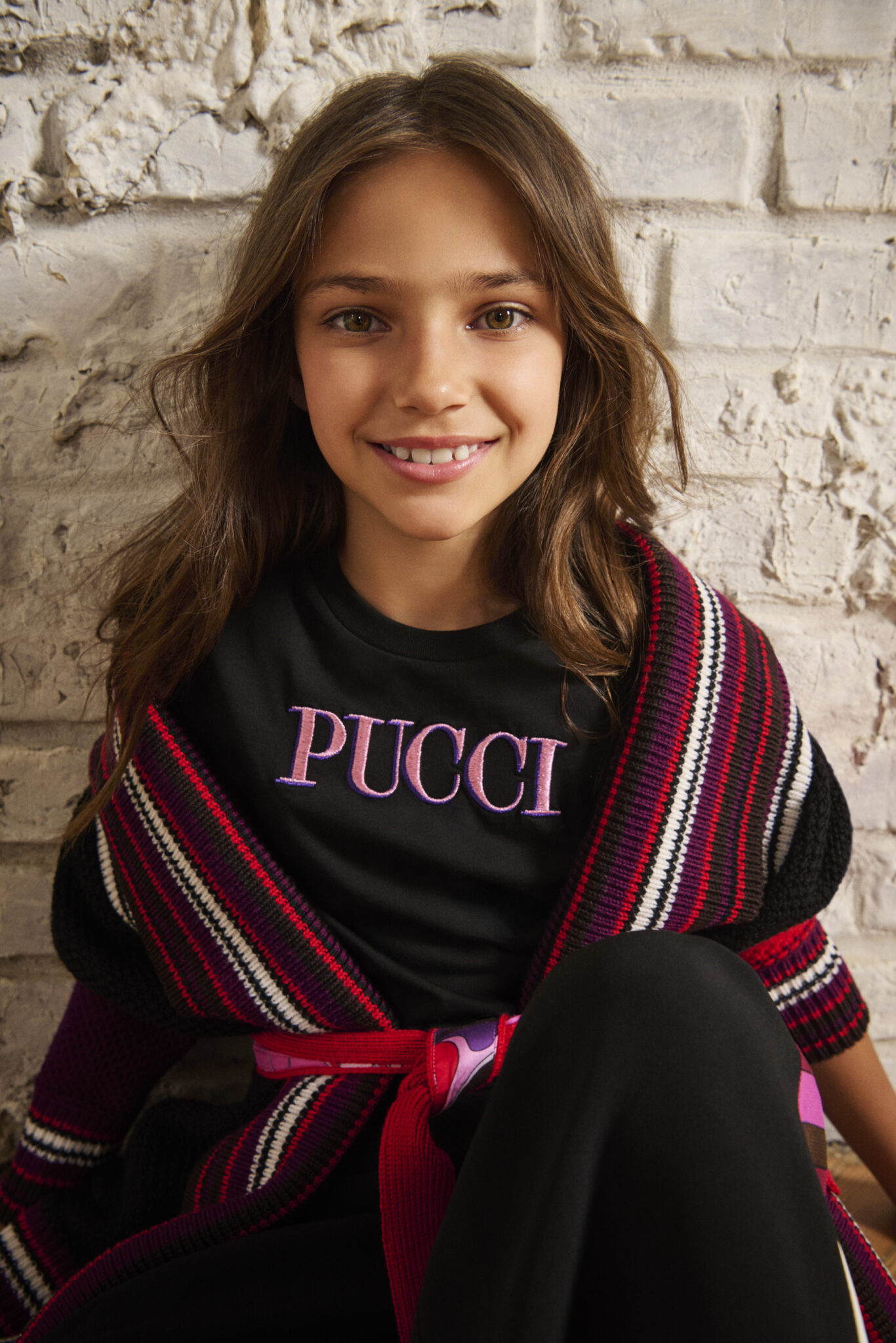 Close-up of a girl smiling, wearing FW25 Pucci Junior striped knit cardigan in red and black, layered over a PUCCI logo T-shirt, featured in Scimparello Magazine.