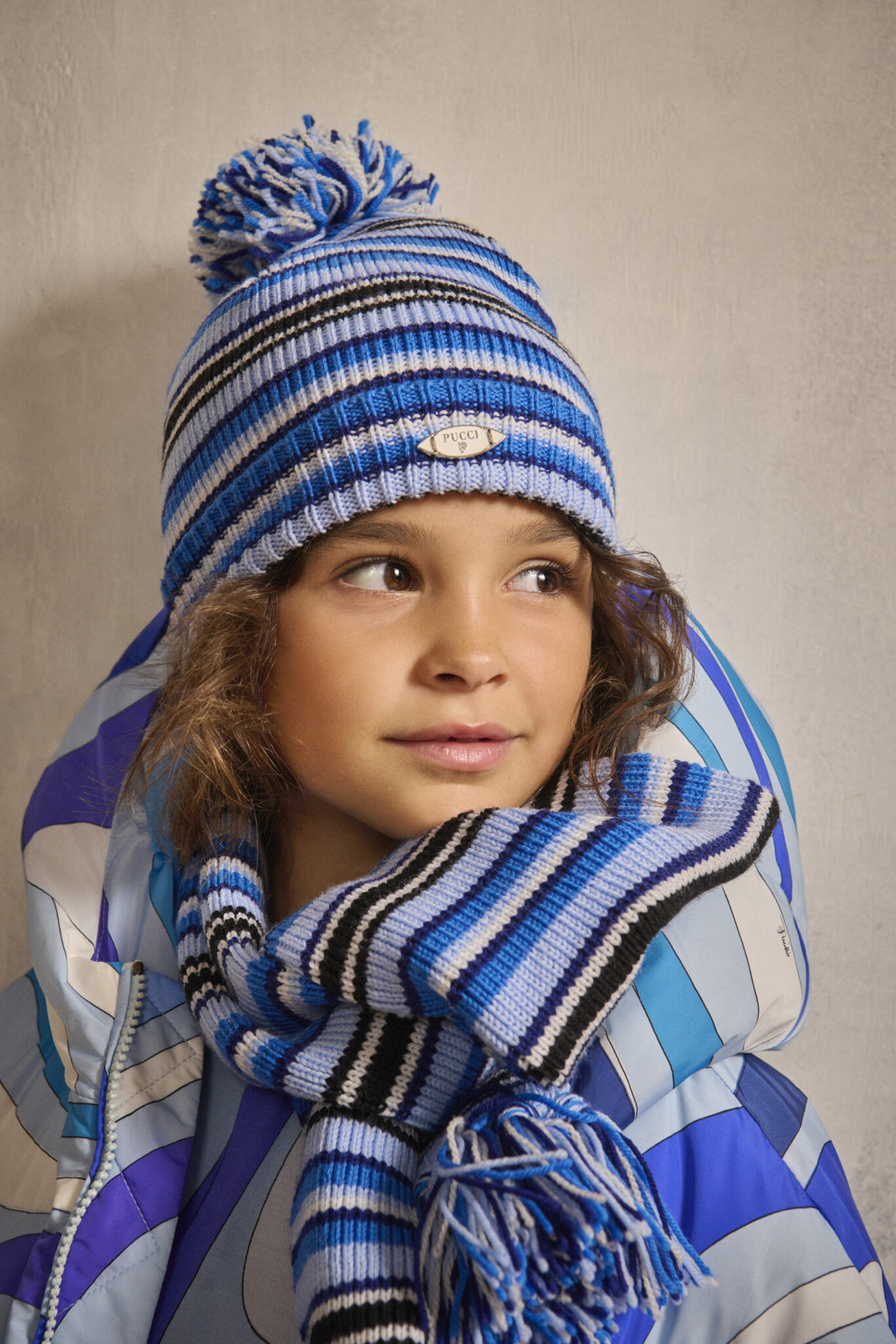 Child wearing FW25 Pucci Junior striped knit hat with pom-pom and matching scarf in shades of blue, paired with a patterned puffer jacket. Photo for Scimparello Magazine.