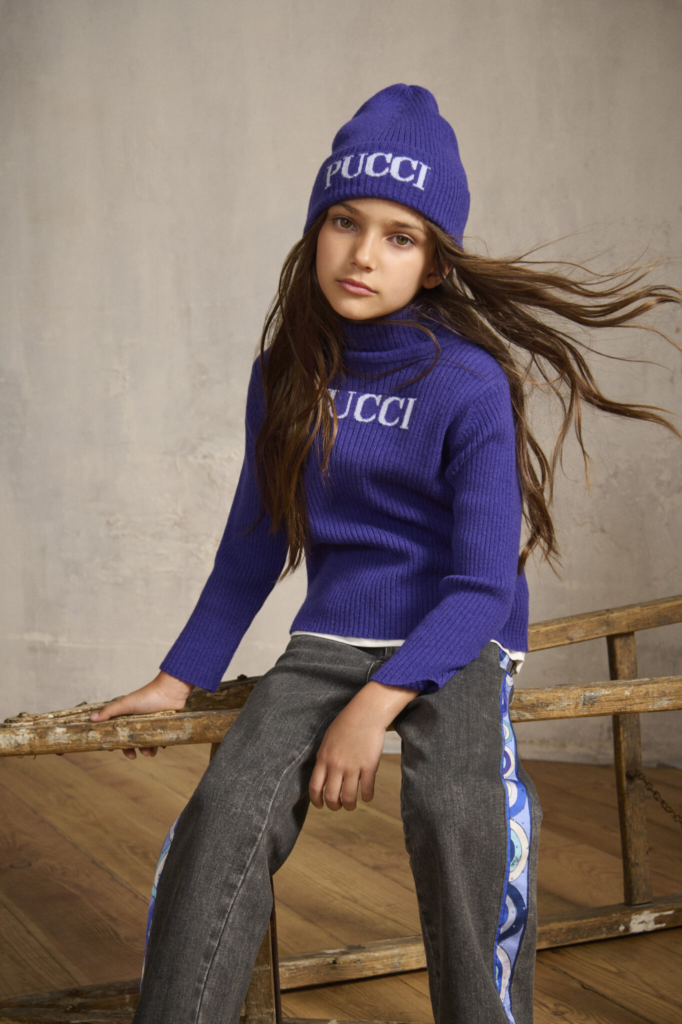 Girl wearing FW25 Pucci Junior purple knit sweater and matching beanie with PUCCI logo, styled with dark trousers featuring printed side bands, featured in Scimparello Magazine.