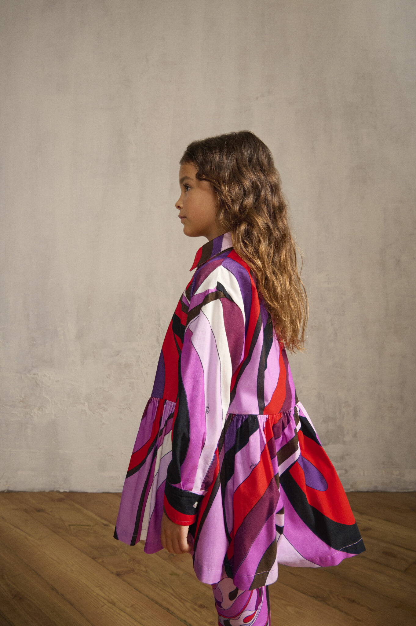 Girl in FW25 Pucci Junior flared shirt dress with bold purple, red, and pink wave print, shown in profile against a neutral background, featured in Scimparello Magazine.