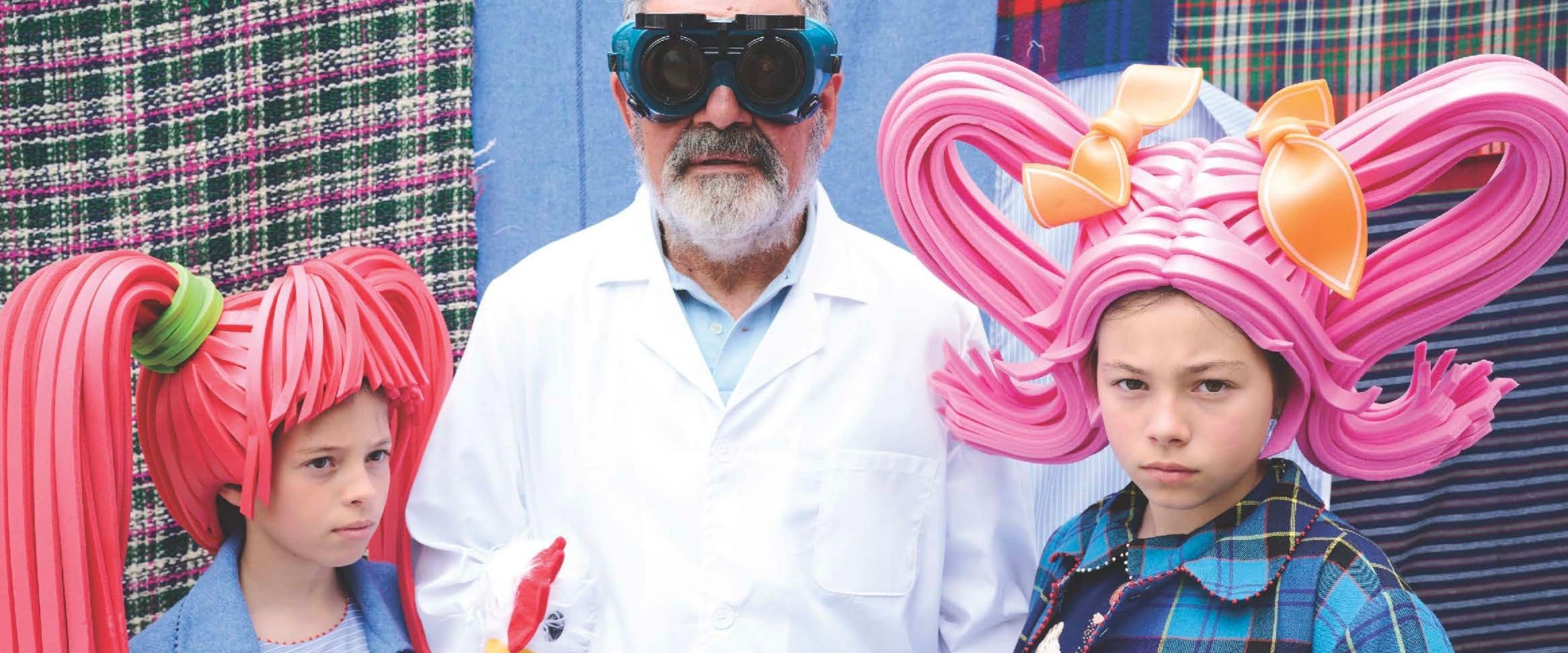 Two children in PÉRO FW25 collection wearing oversized pink foam wigs with bows, standing beside an older man in white coat and goggles, Scimparello Magazine.