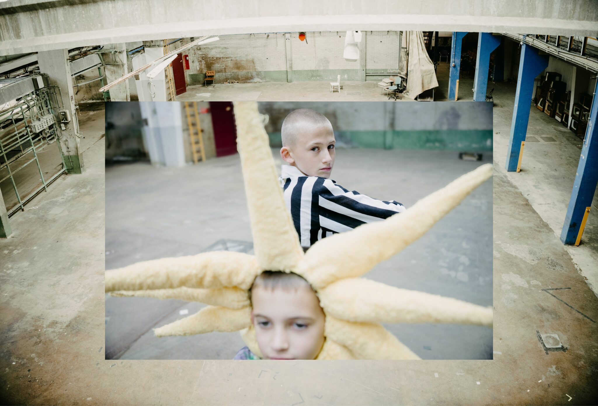 A layered composition inside an industrial warehouse shows two children: in the foreground, a boy wears a plush headpiece shaped like sun rays, while behind him another boy in a bold black-and-white striped jacket looks back over his shoulder with an intense gaze. The background reveals the vast empty warehouse space with exposed beams and painted pillars. Fashion editorial photographed by Aarjan Hamel for Scimparello Magazine.