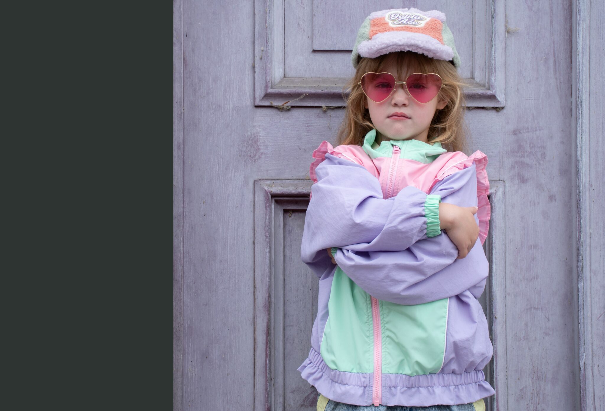 Luisa wearing GOLDIE & ACE FW25 pastel color-block jacket in pink, mint and lilac with ruffle details, GOLDIE & ACE FW25 pants, GOLDIE & ACE FW25 shearling cap, stylist’s own heart-shaped sunglasses and ZARA FW25 shoes, standing in front of a lilac wooden door with arms crossed. Fashion editorial 