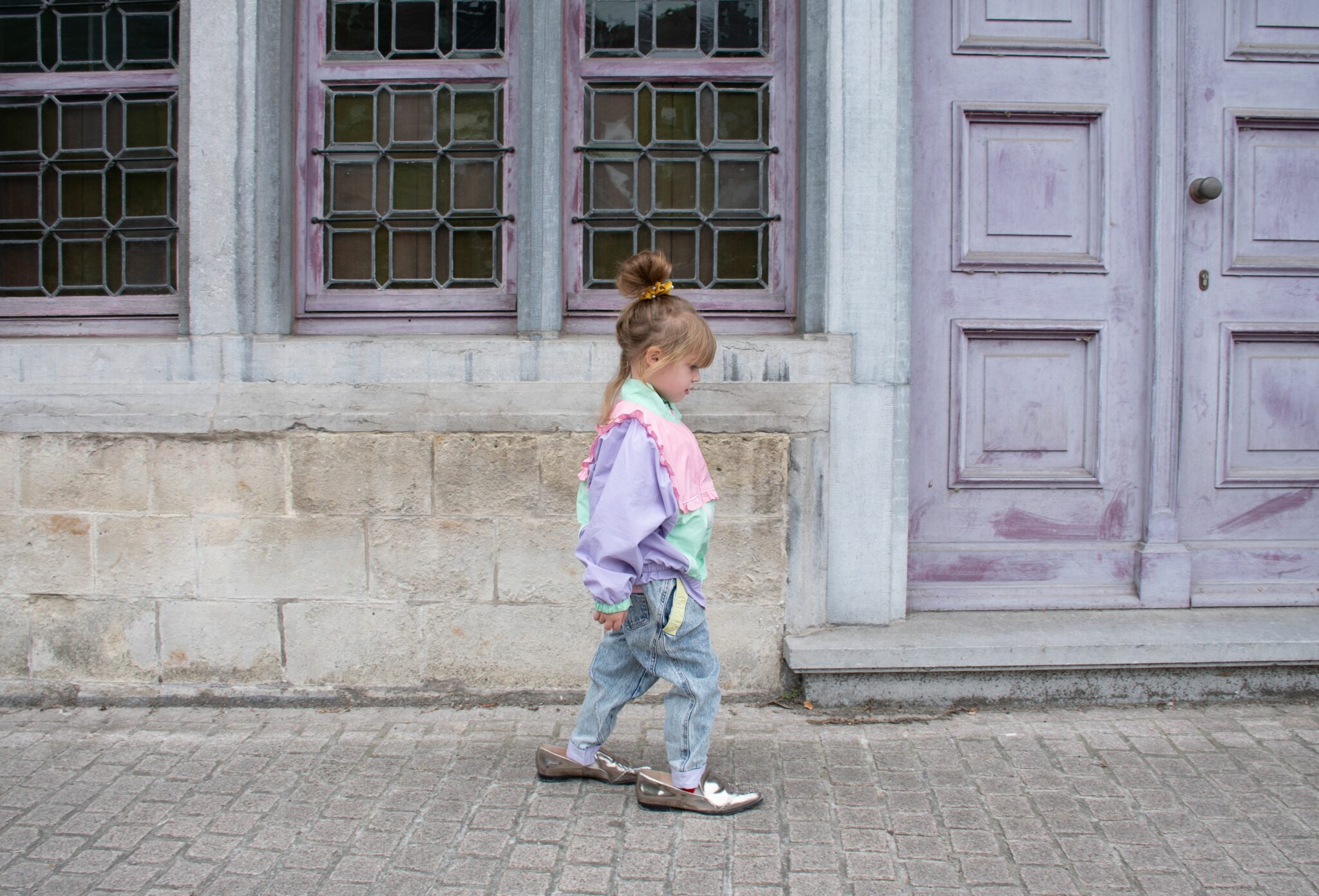 Luisa walking on a cobblestone street in front of lilac wooden doors and vintage windows, wearing GOLDIE & ACE FW25 pastel color-block jacket in pink, mint and lilac with ruffle detail, GOLDIE & ACE FW25 denim pants, ZARA FW25 silver shoes and stylist’s own sunglasses. Fashion editorial 