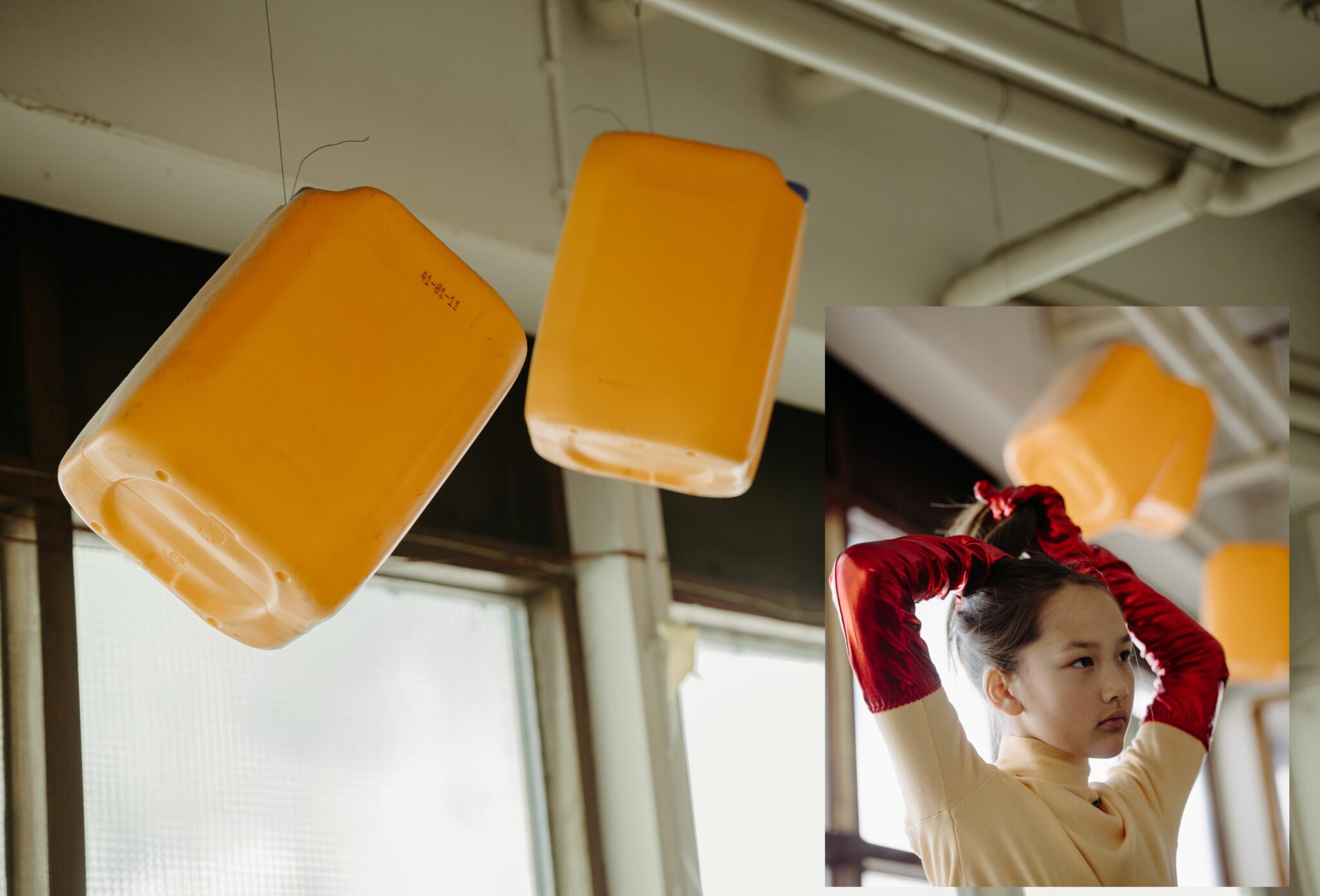 A conceptual diptych: the main image shows bright orange plastic containers hanging from wires against a backdrop of industrial windows and pipes. Overlaid in the foreground, a girl in a cream turtleneck and striking long red satin gloves ties her hair up, with the suspended containers visible in the background. Fashion editorial photographed by Aarjan Hamel for Scimparello Magazine.