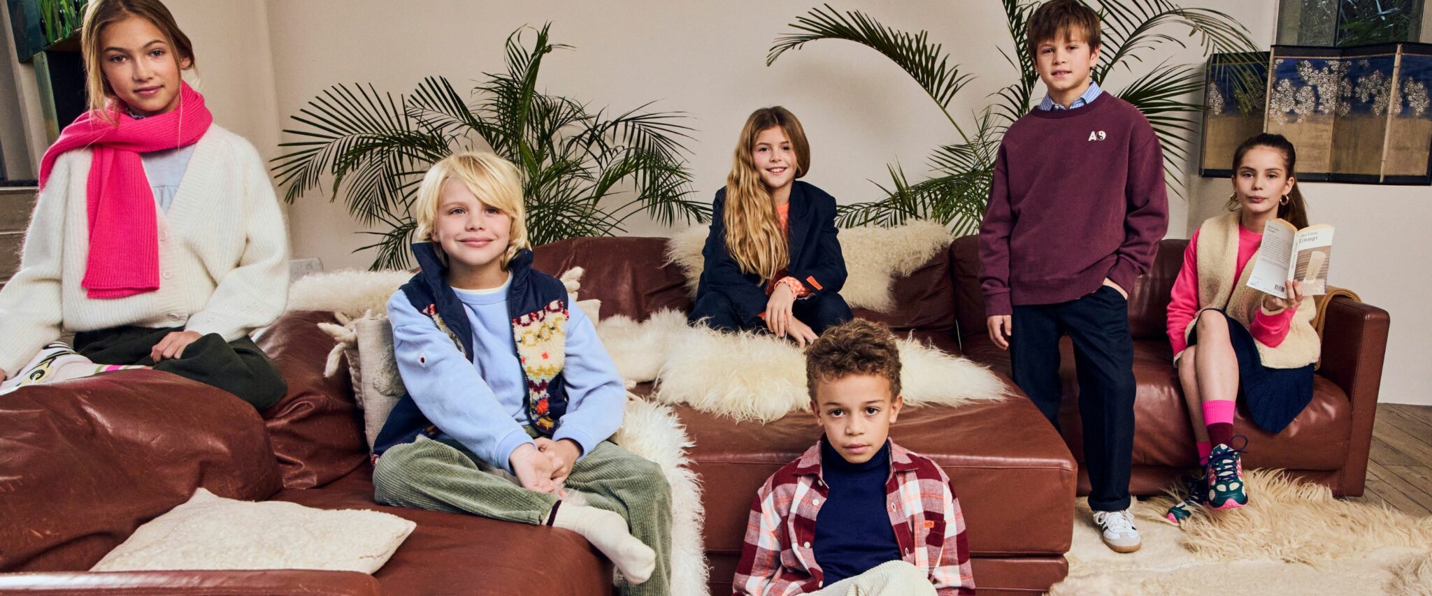 Kids fashion editorial by Scimparello Magazine featuring six children in AO76 Fall Winter 2025 collection. Models wear cozy knit cardigans, colorful scarves, corduroy pants, layered shirts, vests, and sneakers. Styled in a warm living room with leather sofas, wool blankets, and green plants, capturing playful and cozy vibes of kids fashion FW25.
