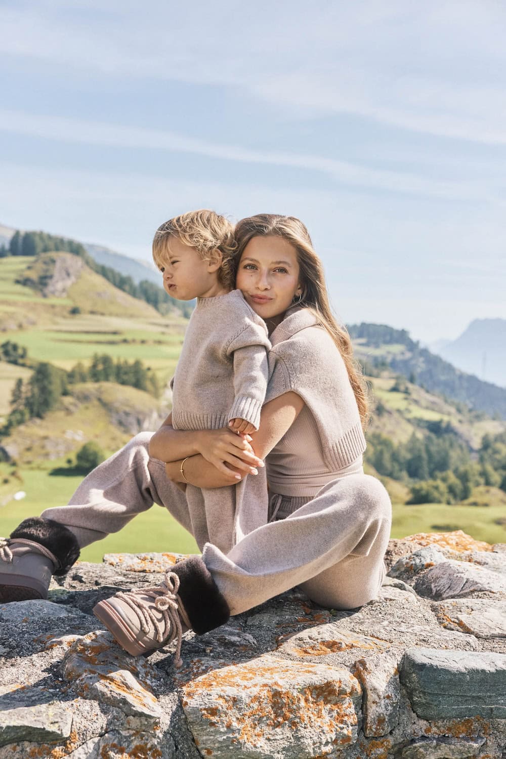 Mother and child in matching beige knitwear from the Minimalisma x Zoë Pastelle capsule collection Fall/Winter 25, sitting outdoors in the mountains, featured on Scimparello Magazine.