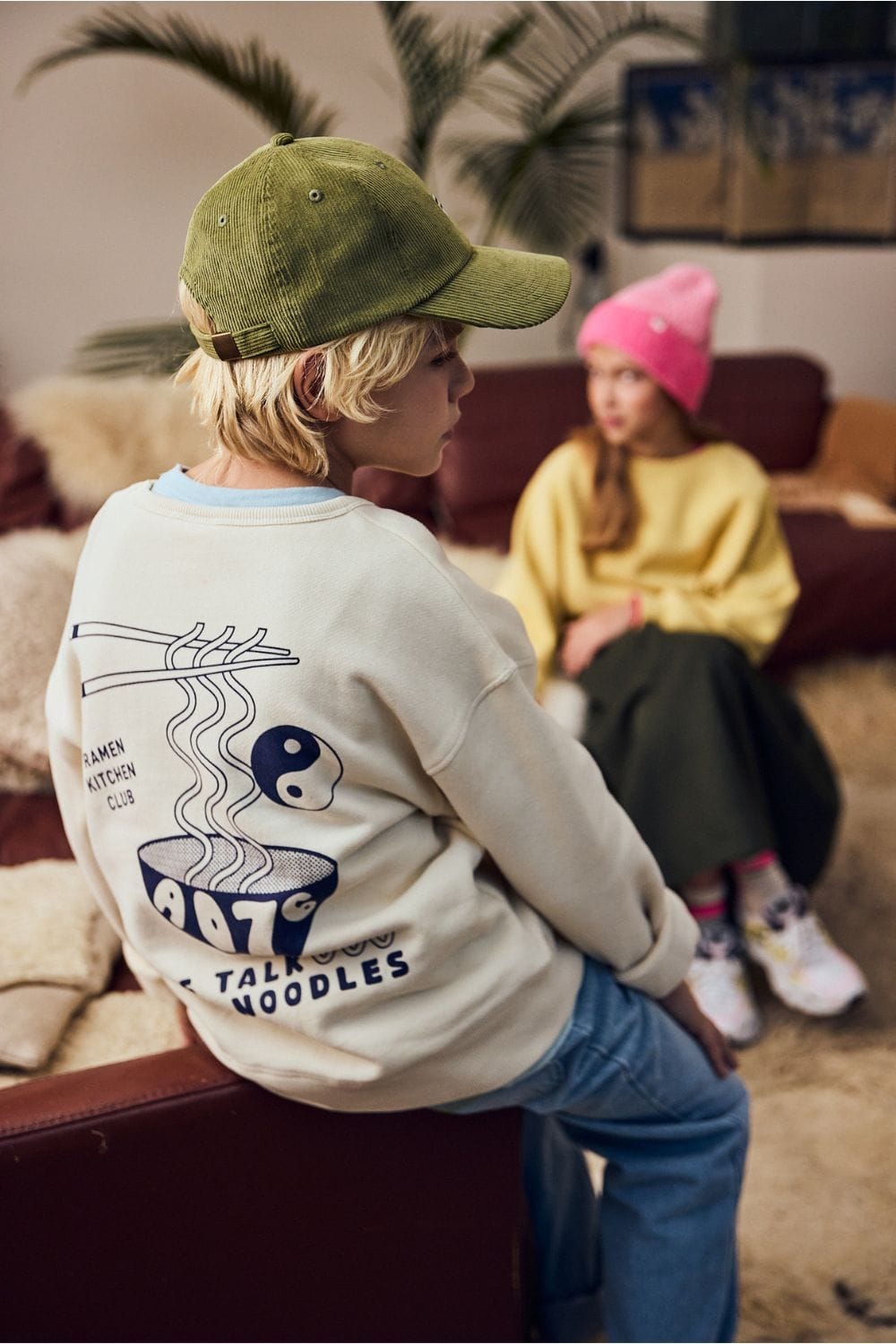 AO76 Kids Collection Fall Winter 2025 on Scimparello Magazine. Six children in cozy layered outfits with knit cardigans, scarves, corduroy pants, vests and sneakers, sitting on brown leather sofas with wool blankets and green plants in the background. Playful and stylish kids fashion FW25.