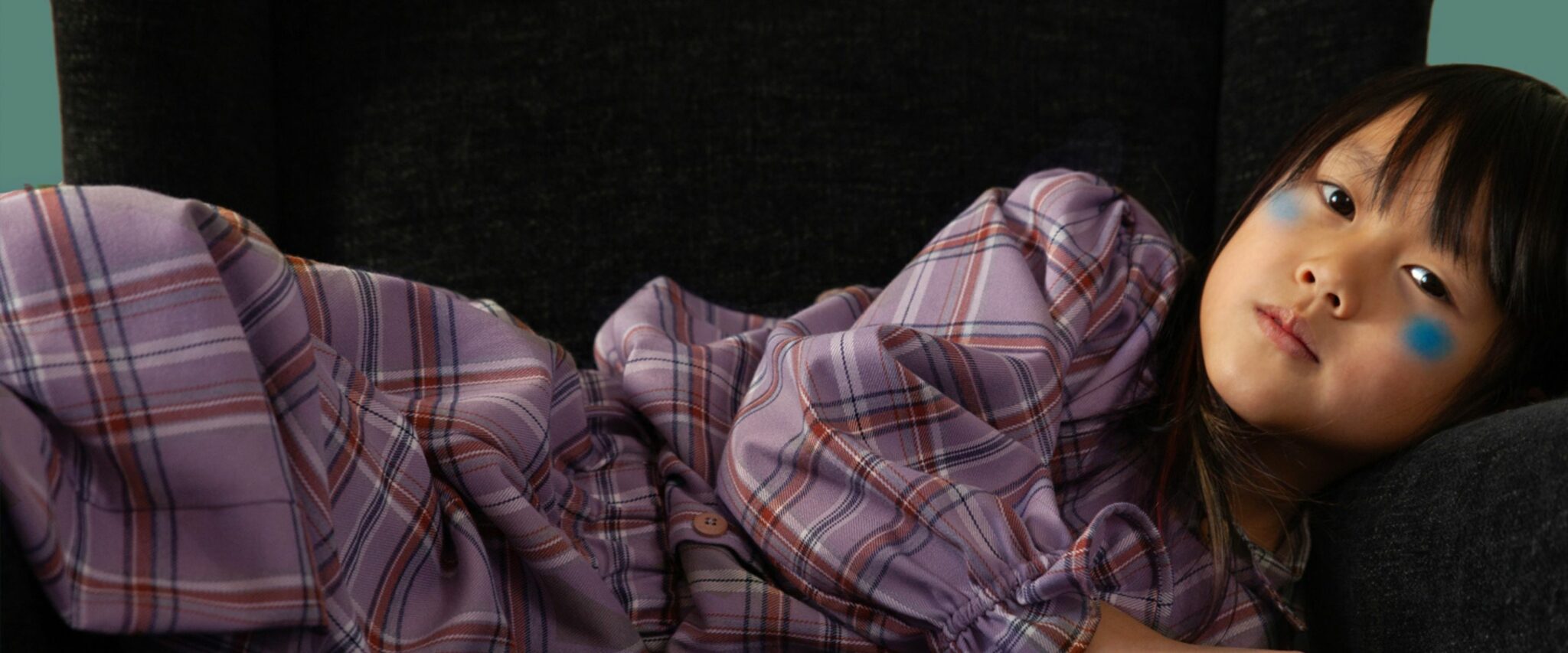 Child reclining on a dark armchair wearing a purple checkered dress from Bianca & Noè FW25, featured in Scimparello Magazine, with blue painted cheeks adding a playful touch.