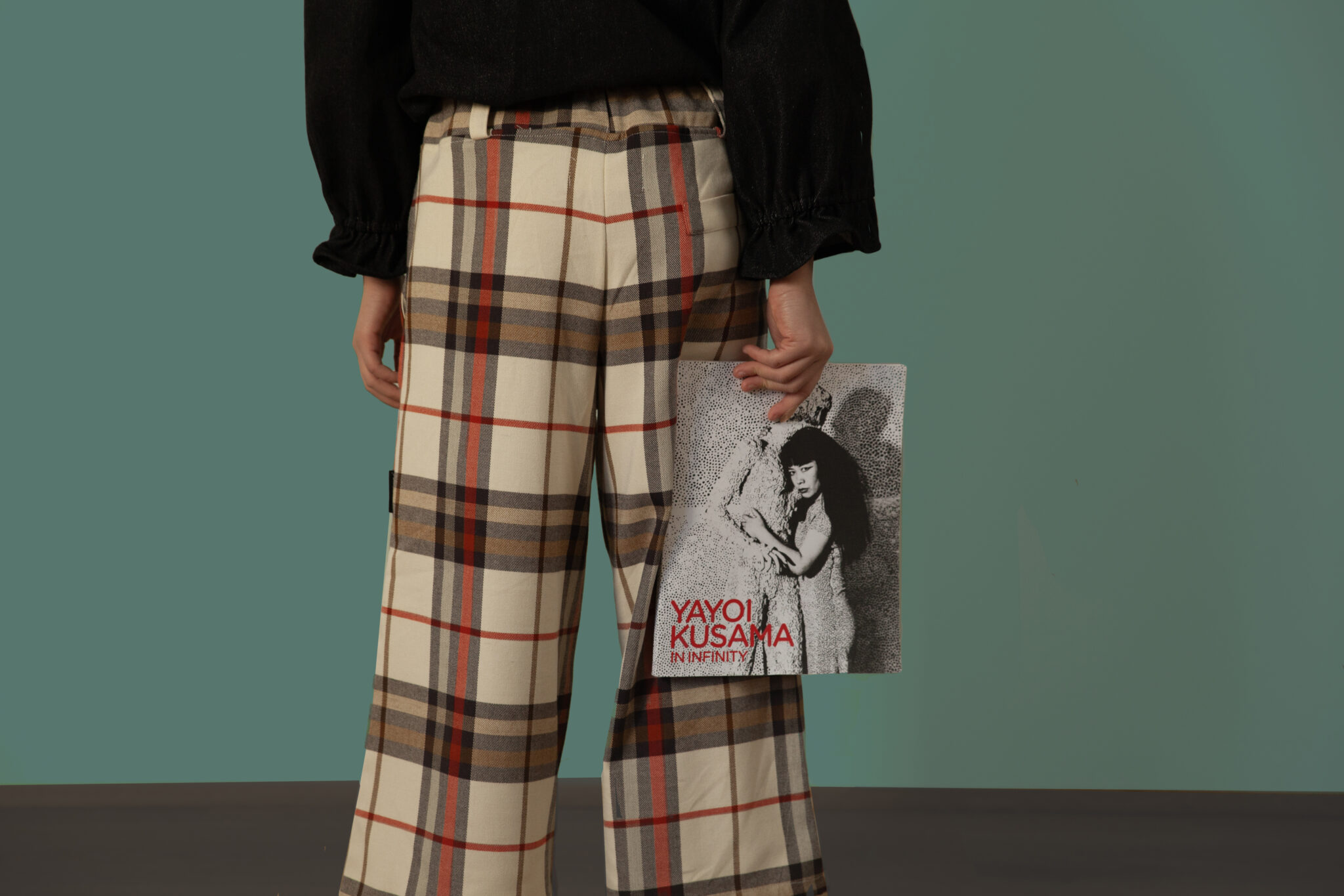 Close-up of a child from the Bianca & Noè FW25 kidswear collection for Scimparello Magazine, showing the back of beige plaid trousers with red and brown checks. They hold a book titled Yayoi Kusama: In Infinity in one hand, paired with a black shirt. Children’s fashion FW25.