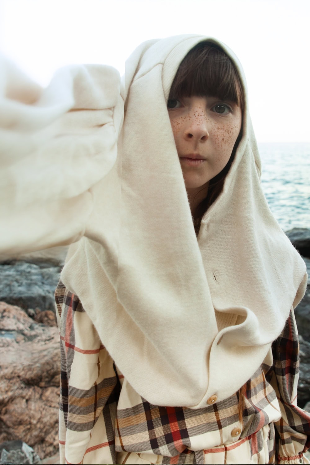 A child stands by the seaside rocks, wearing a cream hooded piece draped over the head and a plaid button-up dress in earthy tones of beige, brown, and red. The styling feels natural and raw, highlighting the textures of fabric against the ocean backdrop. Fashion editorial FW25 Bianca & Noè for Scimparello Magazine.