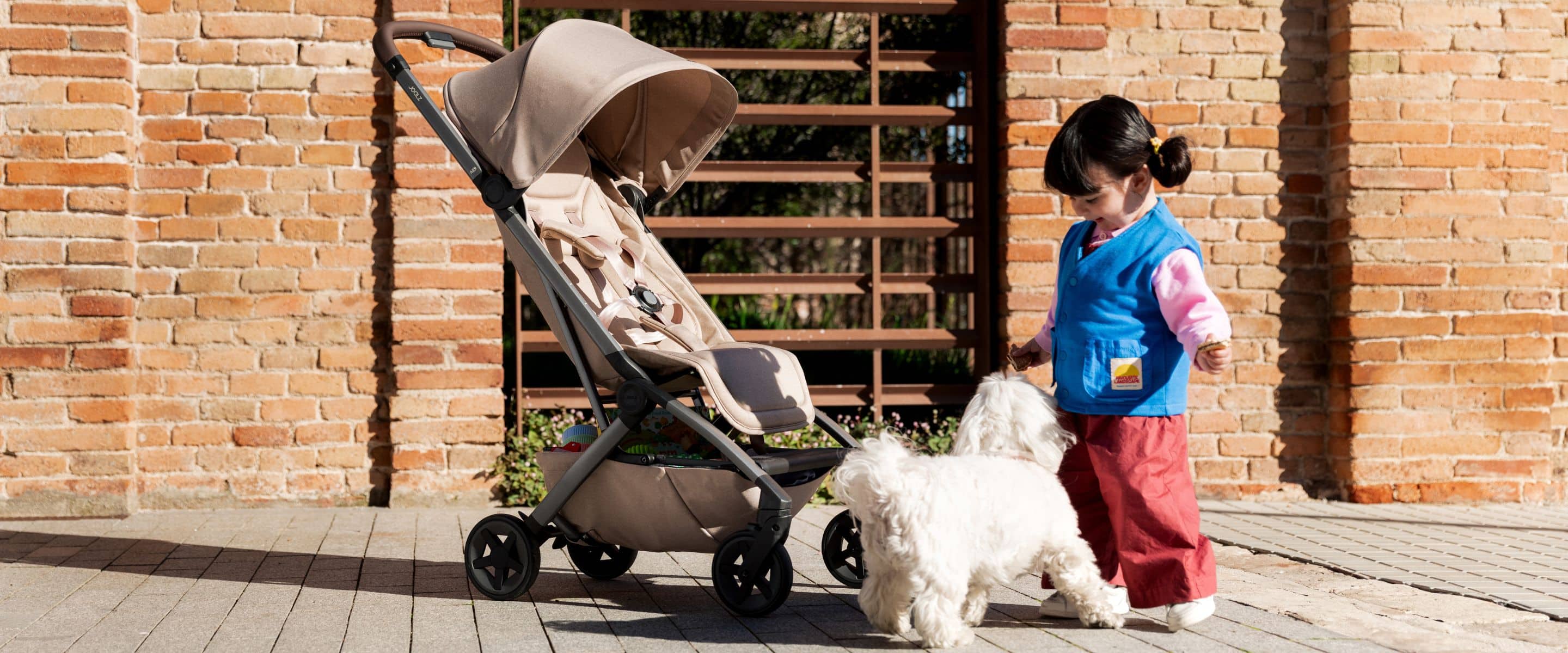 Joolz stroller collection – a beige compact stroller next to a little girl playing with a white dog, outdoor urban setting.