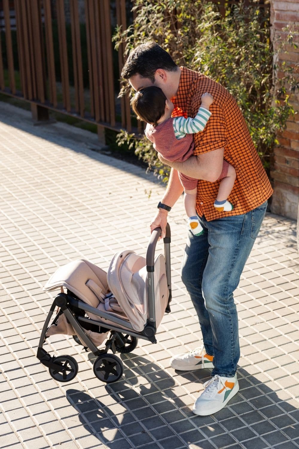 Father holding baby while folding a beige Joolz stroller outdoors, showing the compact and practical design, part of the Joolz stroller collection.