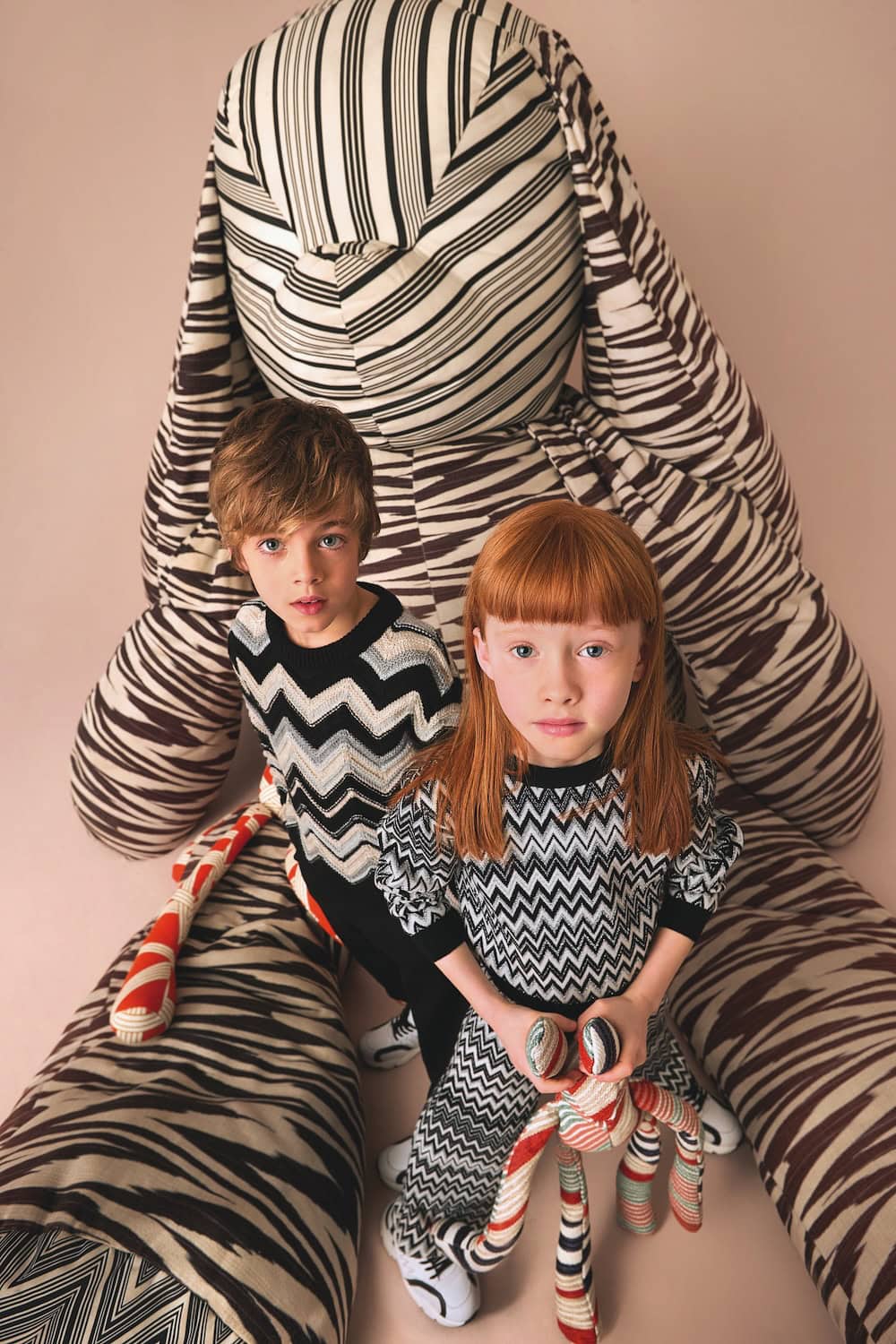 Two children wearing FW25 Missoni Kids zigzag patterned knitwear in black and white tones, posing in front of an oversized striped plush bunny. Photo for Scimparello Magazine.