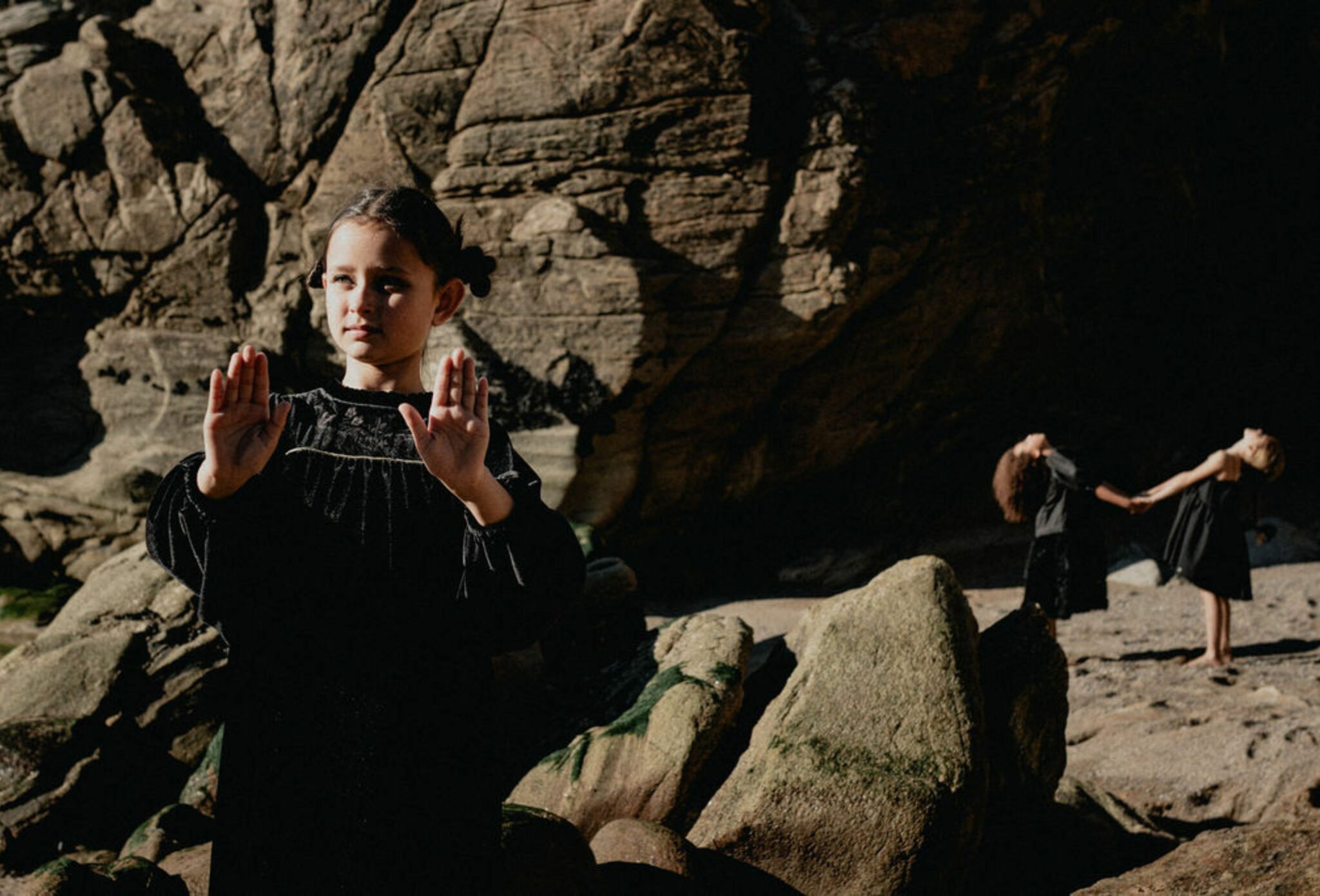 Three girls dressed in black perform a quiet, mystical ritual among seaside rocks — one stands in the foreground with raised hands as if casting a spell, while the other two hold hands and lean back in unison under the sunlight. From the FW25 fashion editorial Coven III – Black Witches by photographer Lara Lutz for Scimparello Magazine, celebrating magic, nature, and sisterhood.