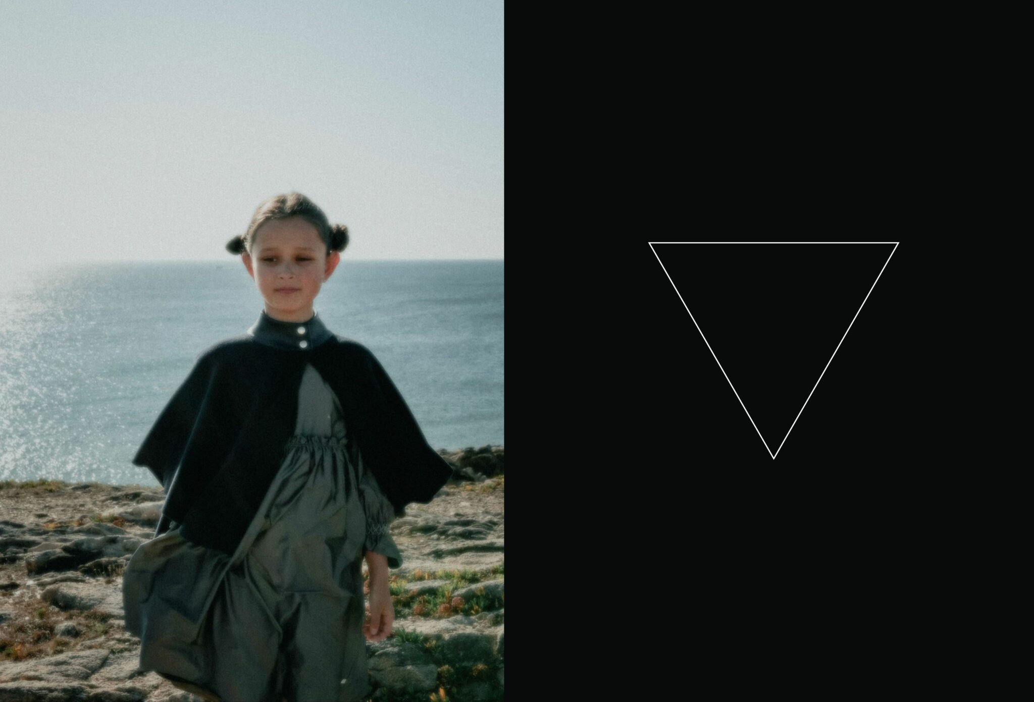 A girl dressed in a dark cape and grey dress walks along a rocky coastline under soft light, the sea shimmering behind her. Beside her, a minimalist black panel features a thin inverted triangle — a recurring symbol of the Coven III – Black Witches FW25 editorial by photographer Lara Lutz for Scimparello Magazine, evoking mystery, balance, and transformation.