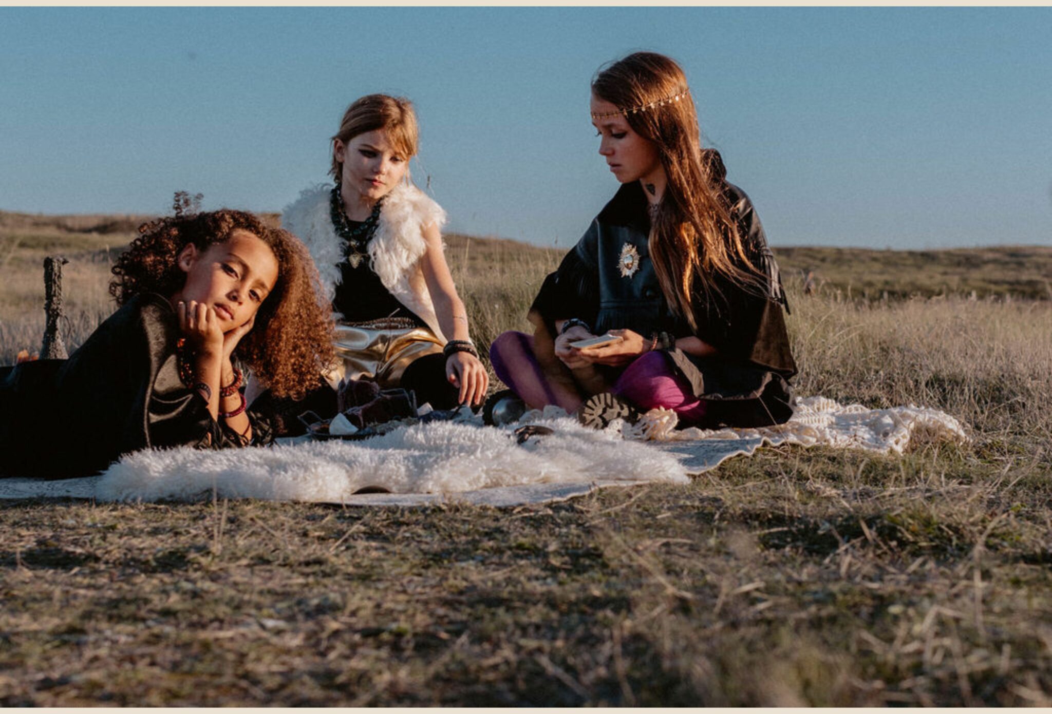 Three girls sit on a blanket in a sunlit field, surrounded by tarot cards and bohemian accessories — one in a faux-fur vest, one with golden pants, and one wearing a headpiece and holding the cards. From Coven IV – Gipsy Rock & Tarot Magic by Lara Lutz for Scimparello Magazine, this Halloween-themed kids fashion editorial mixes rock spirit, mystical energy, and boho charm.
