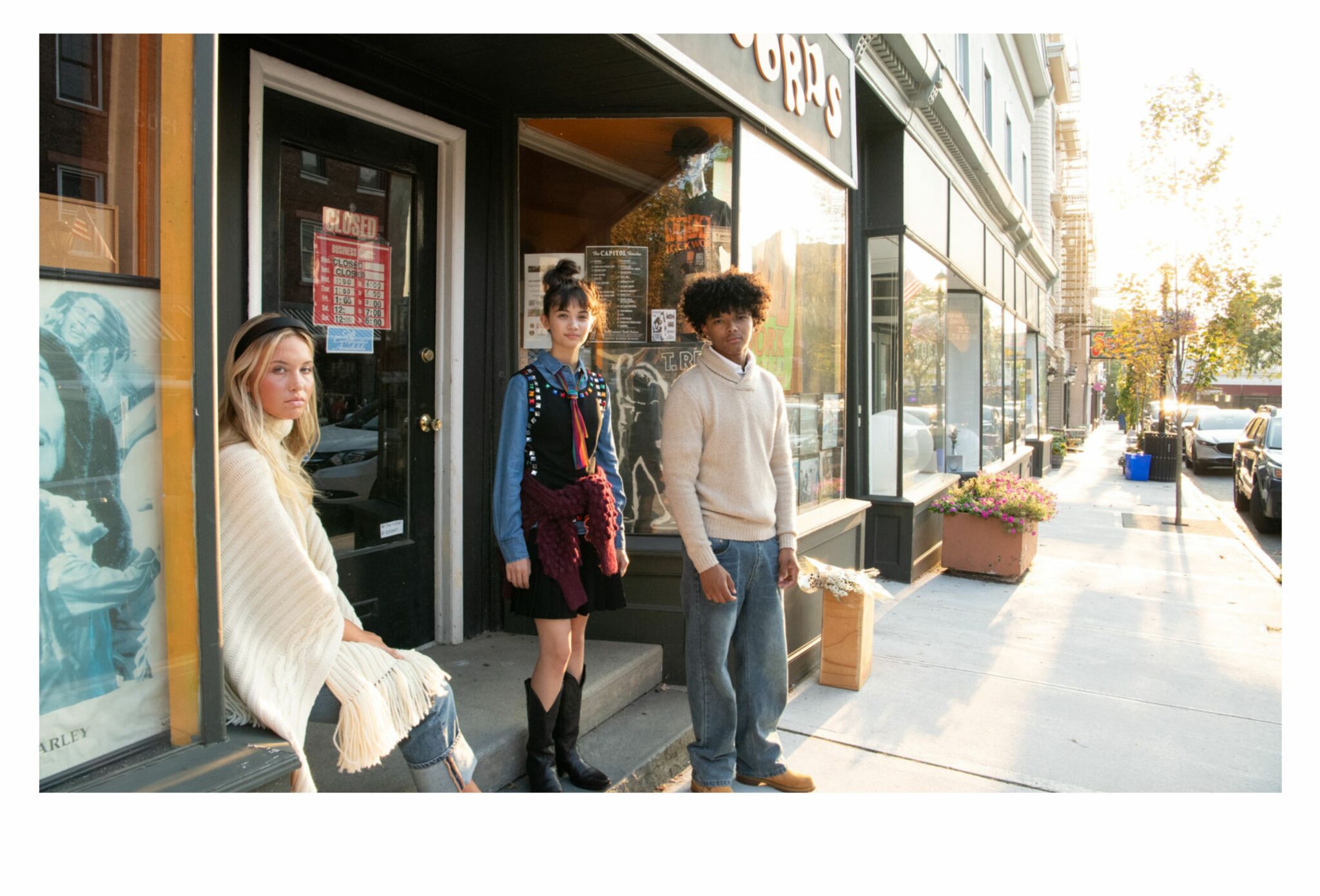 Three teens in fall outfits standing outside a record shop at sunset — one wearing a cream fringed poncho, one in a denim shirt layered under a studded black dress with a red sweater tied at the waist, and one in a beige turtleneck sweater with jeans. The golden light reflects off the shop windows, creating a warm nostalgic mood. Teen fashion editorial All the Time in the World by Sabrina Helas for Scimparello Magazine FW25.