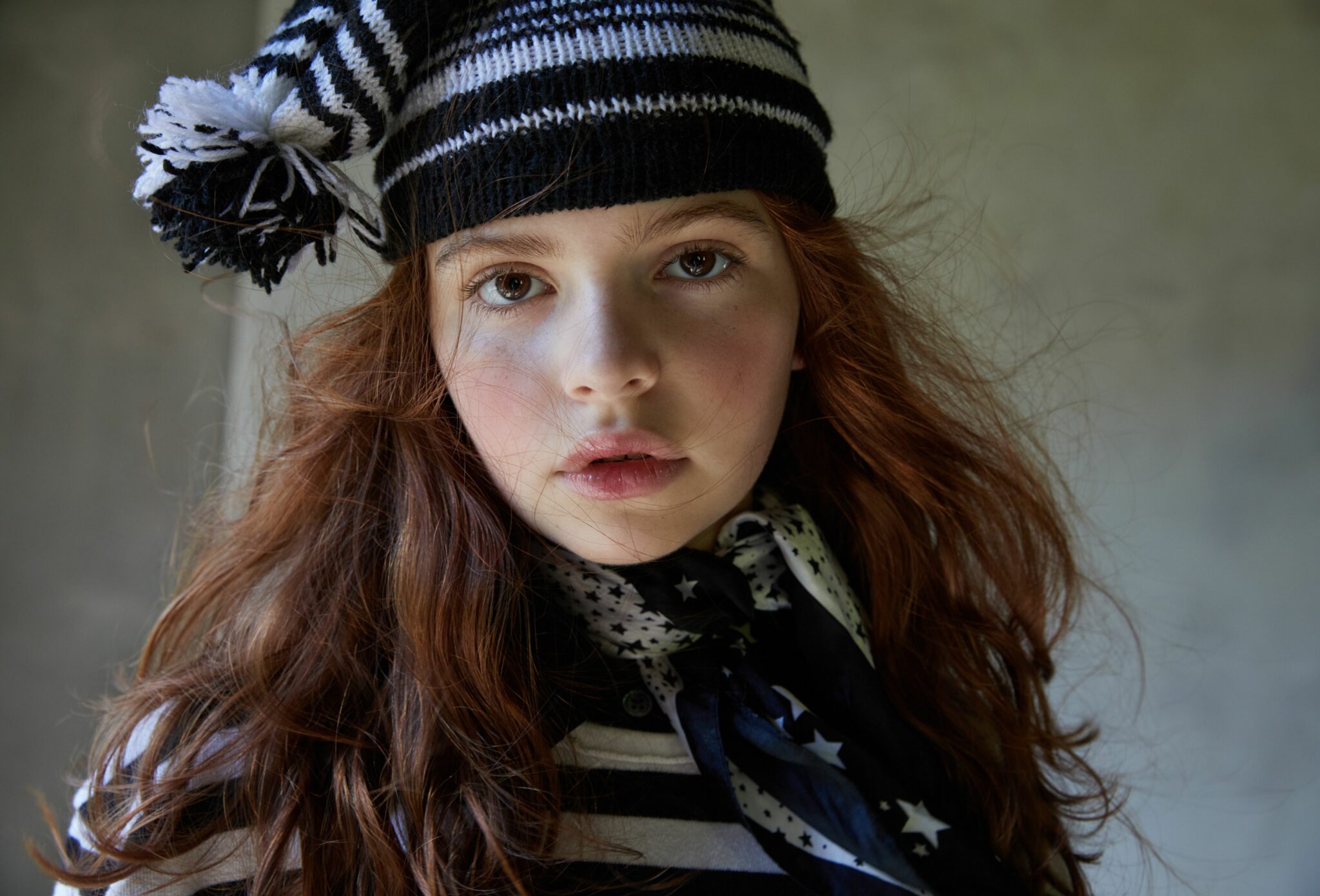 Fashion editorial That’s Bonkers for Scimparello Magazine FW25, photographed by Cleo Sullivan. Close-up portrait of a red-haired girl wearing a black-and-white striped knit hat by IGBO FW25, star-print scarf ADRIENNE VITTADINI FW25, and striped t-shirt JOE’S FW25. The look mixes playful patterns with soft natural light for a poetic yet bold atmosphere.