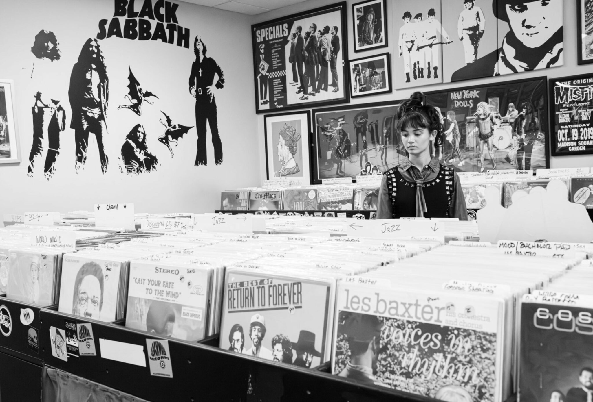 Teen girl browsing vinyl records in a vintage record shop decorated with posters of Black Sabbath, The Specials, and New York Dolls. She wears a denim shirt layered under a studded black vest with a ribbon tie, creating a nostalgic rock-inspired look. Teen fashion editorial All the Time in the World by Sabrina Helas for Scimparello Magazine FW25.