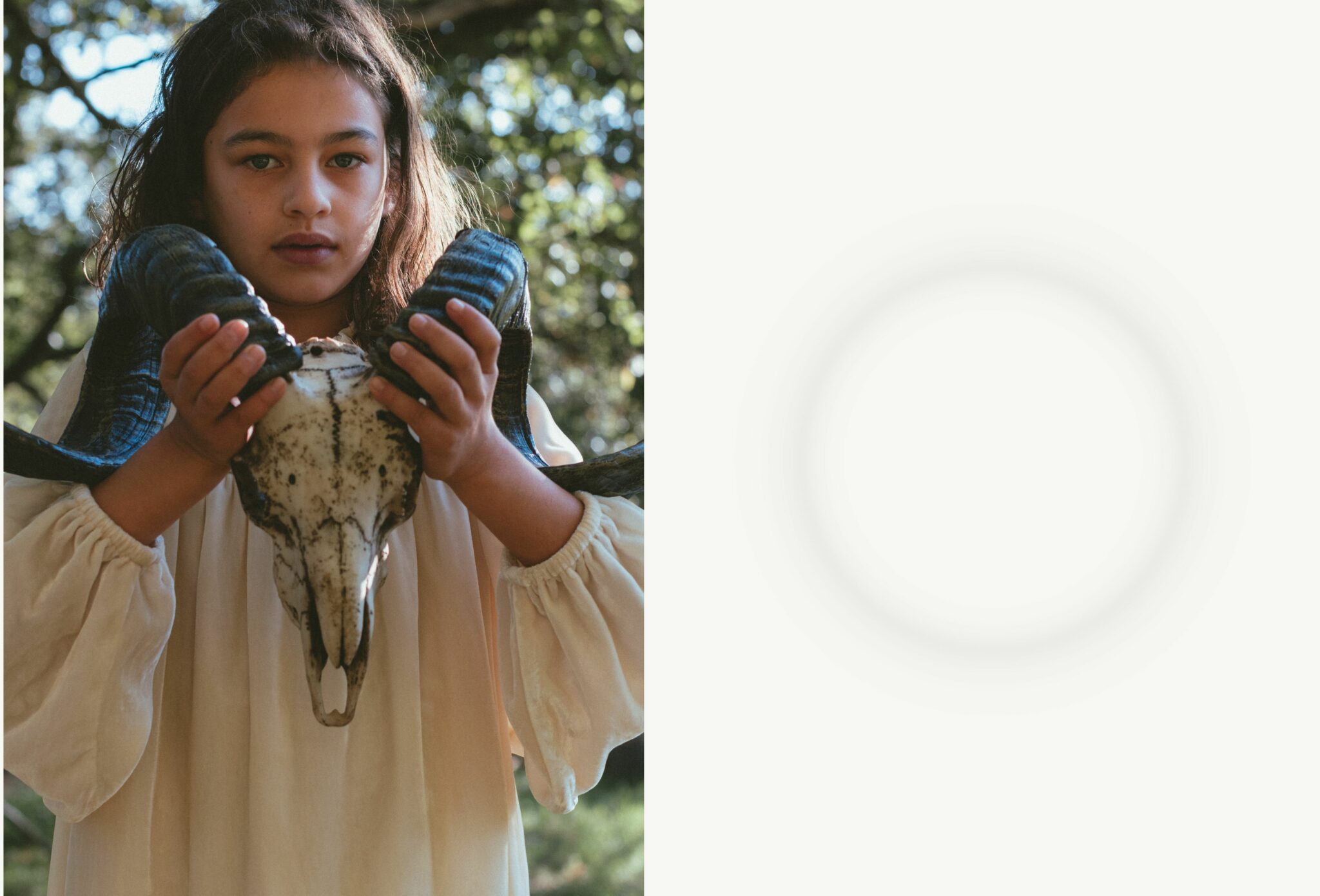 Child in a cream dress holds a large ram skull under dappled sunlight, evoking ritual and mystery. Kids fashion editorial COVEN I by Lara Lutz for Scimparello Magazine, inspired by Halloween and folklore.