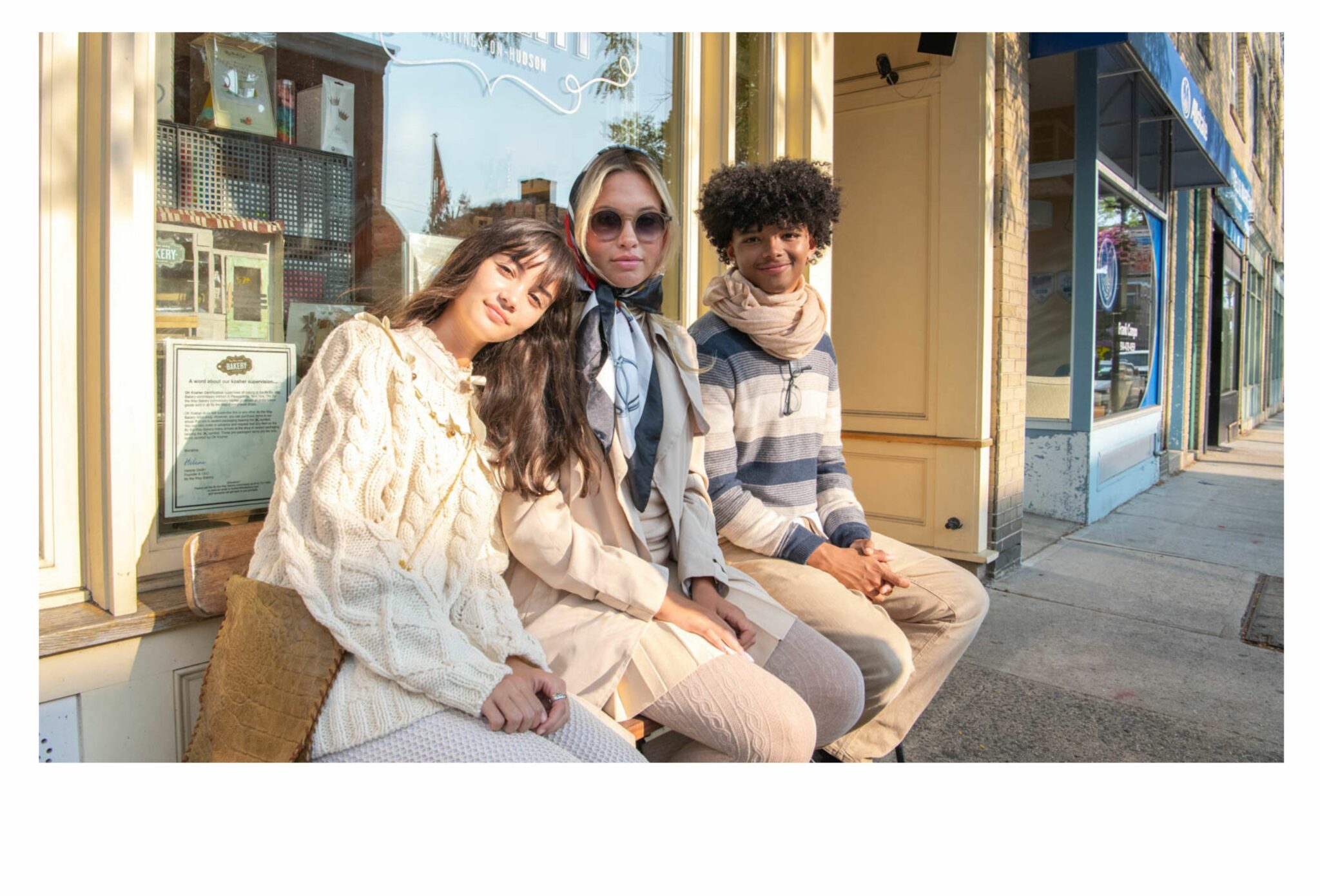 Three teens sitting outside a café in warm afternoon light — one wearing a chunky cream knit sweater, one in a beige trench coat with a silk scarf and sunglasses, and one in a striped pullover with a scarf around the neck. Their relaxed mood and golden glow capture an autumn vibe. Teen fashion editorial All the Time in the World by Sabrina Helas for Scimparello Magazine FW25.