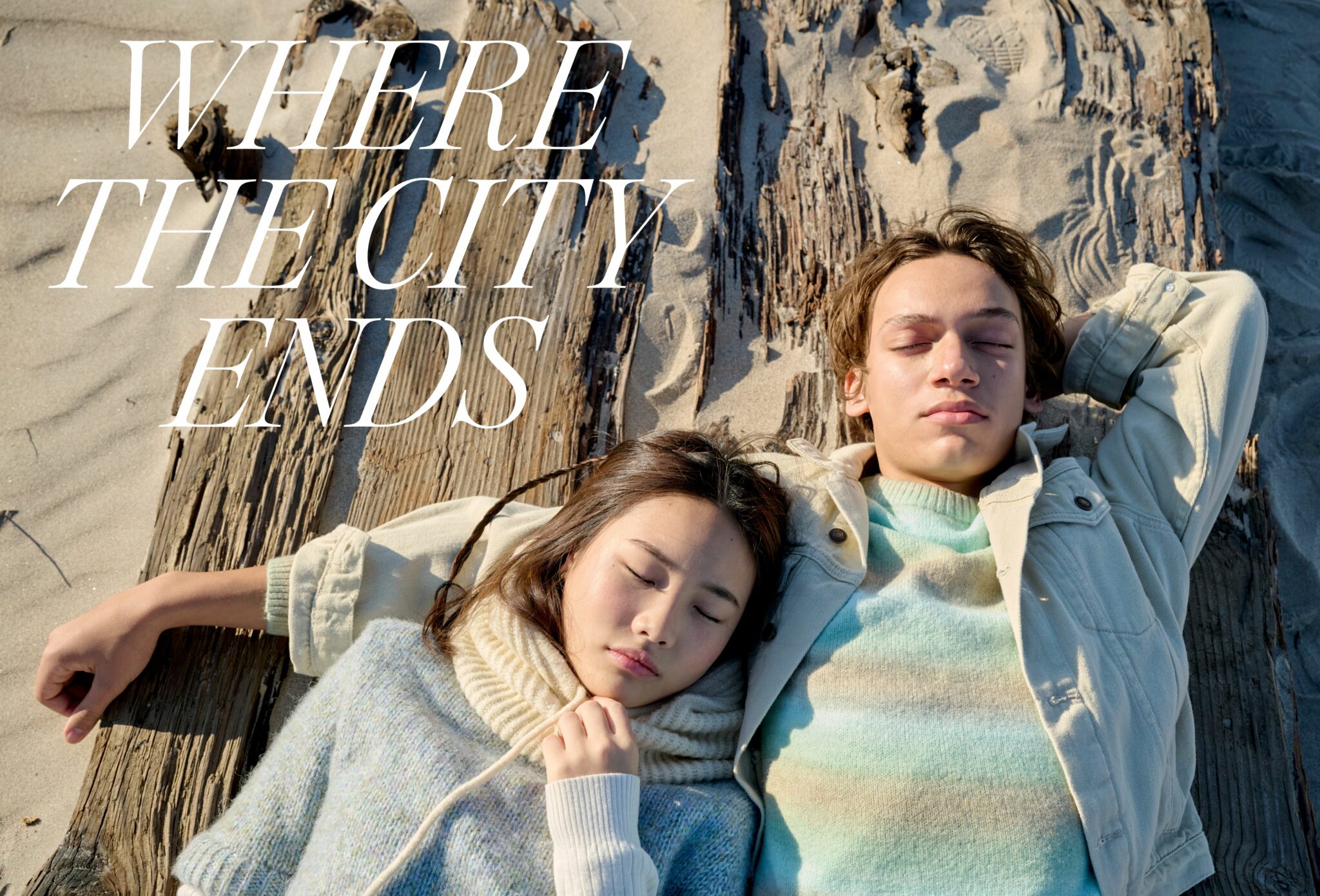 Two teenagers rest on a sandy beach against weathered wooden planks, eyes closed under the sun. The girl wears a soft knitted turtleneck in pale tones, and the boy wears a light jacket over a pastel striped sweater. A quiet, natural moment captured where the city fades into nature. Photo by Jay Fenwick, editorial published in Scimparello Magazine.