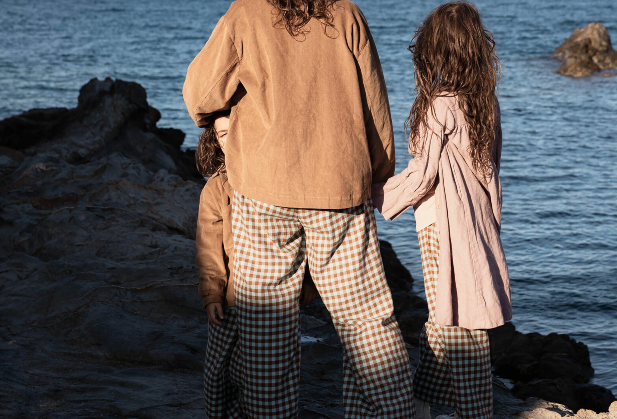 Three children standing on coastal rocks wearing checked trousers and oversized jackets, facing the sea in warm afternoon light. Photo by Manuela Franjou for Scimparello Magazine.
