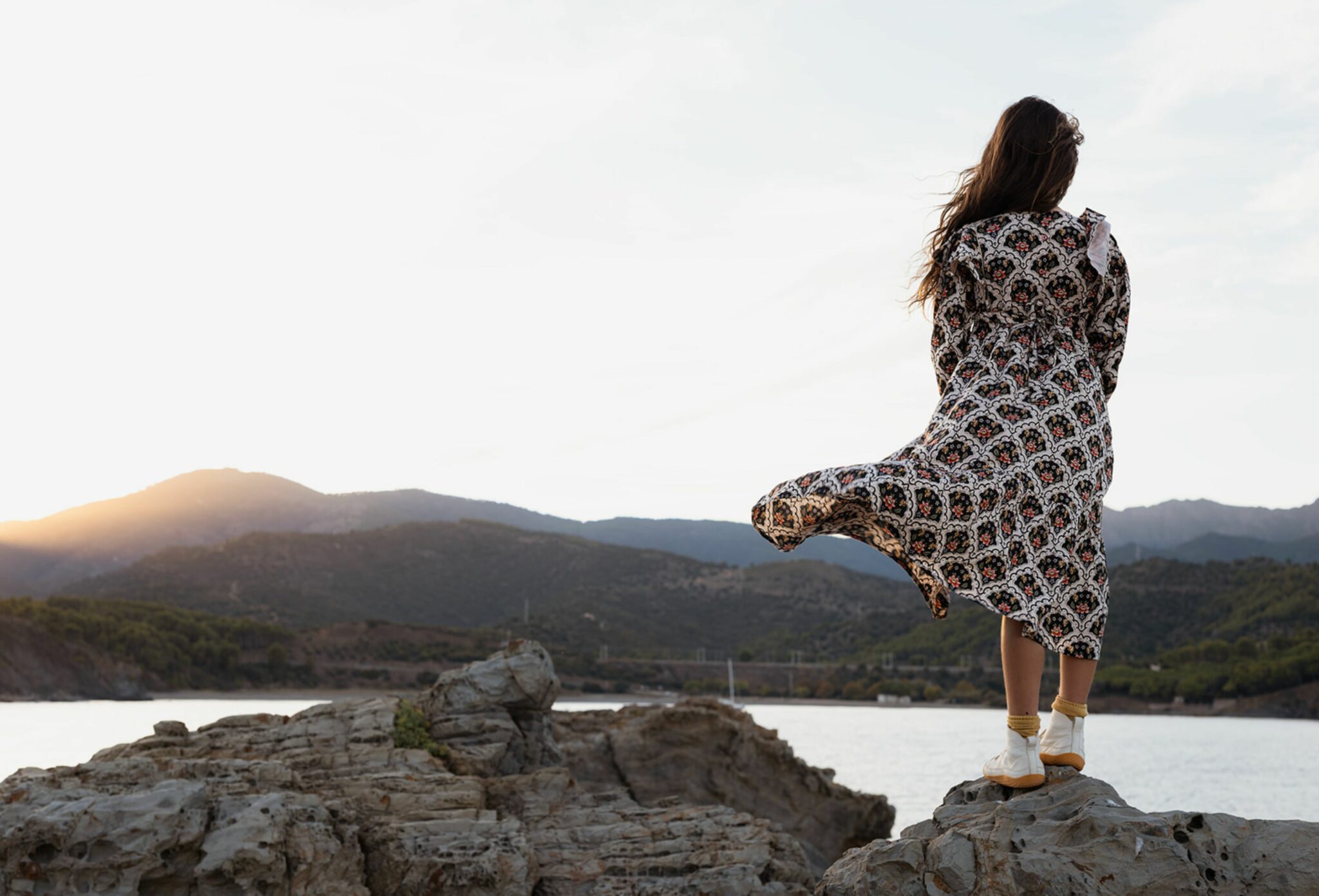 Girl standing on coastal rocks at sunset, wearing a long patterned dress that blows in the wind while she looks toward the mountains and the sea. Photo by Manuela Franjou for Scimparello Magazine.