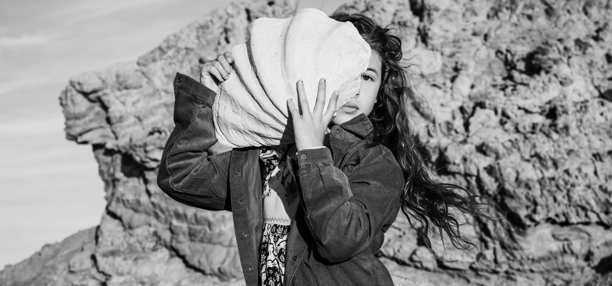 Girl holding a large shell in front of her face in a rocky landscape, photographed by Manuela Franjou for Scimparello Magazine.