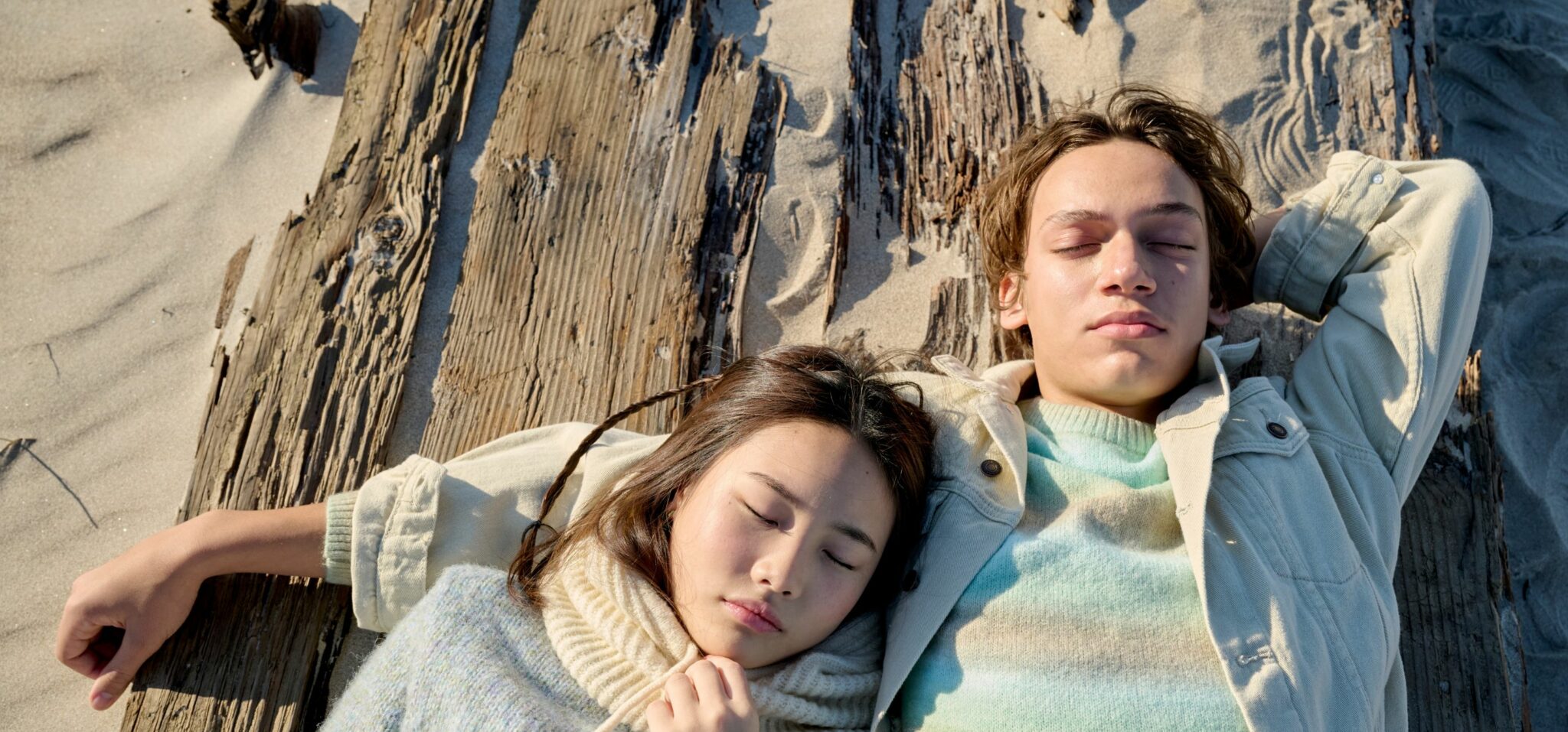 Two teenagers rest on a sandy beach against weathered wooden planks, eyes closed under the sun. The girl wears a soft knitted turtleneck in pale tones, and the boy wears a light jacket over a pastel striped sweater. A quiet, natural moment captured where the city fades into nature. Photo by Jay Fenwick, editorial published in Scimparello Magazine.