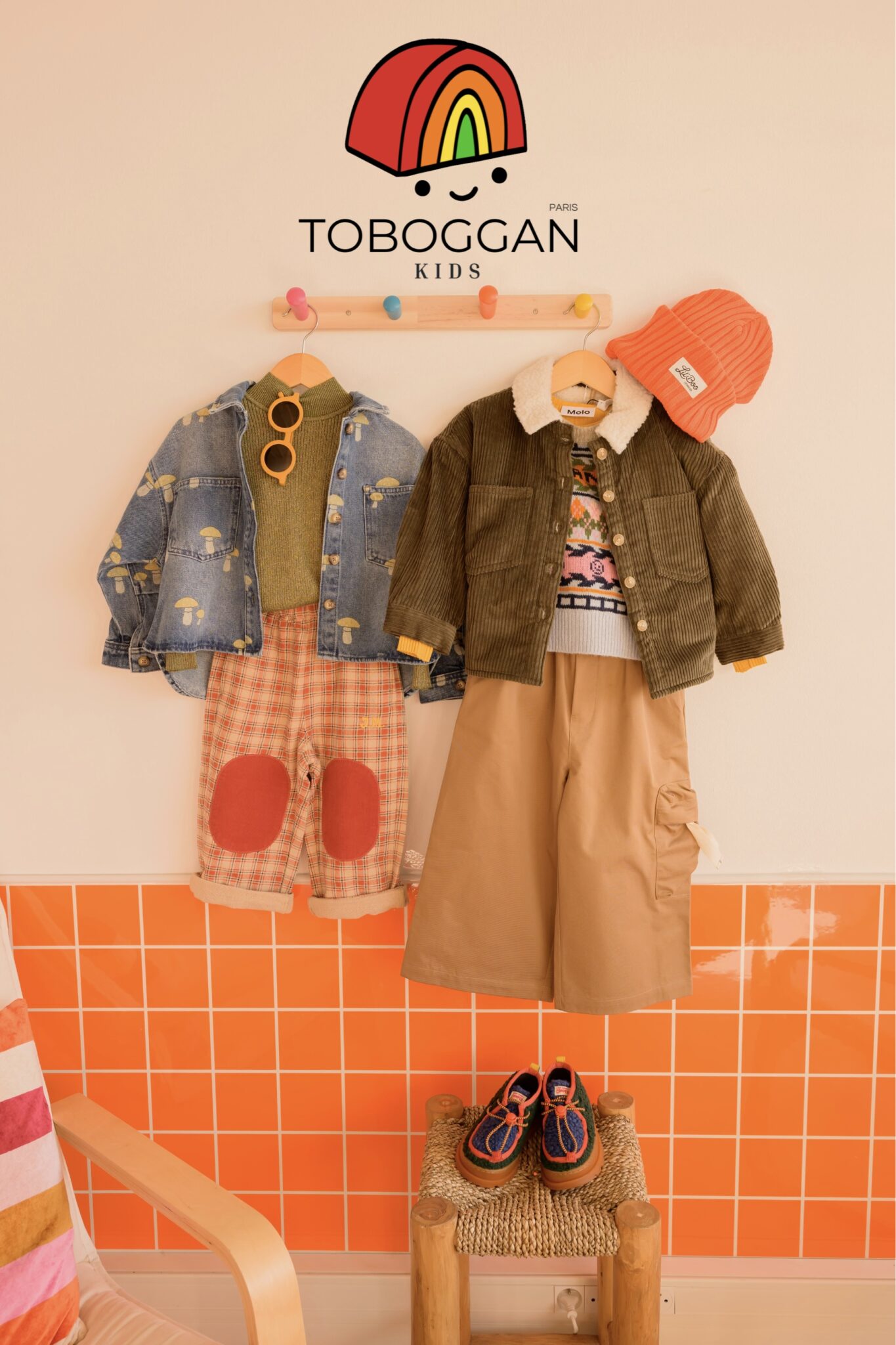 Two colorful kids’ outfits displayed on a wall under the TOBOGGAN KIDS Paris logo, featured on Scimparello Magazine. The left look includes a mushroom-print denim jacket, green knit, and checked pants with red patches; the right look includes a corduroy jacket with shearling collar, graphic knit, wide beige pants, orange beanie, and matching shoes. A warm, retro-inspired styling scene from the online shop Toboggan Kids.