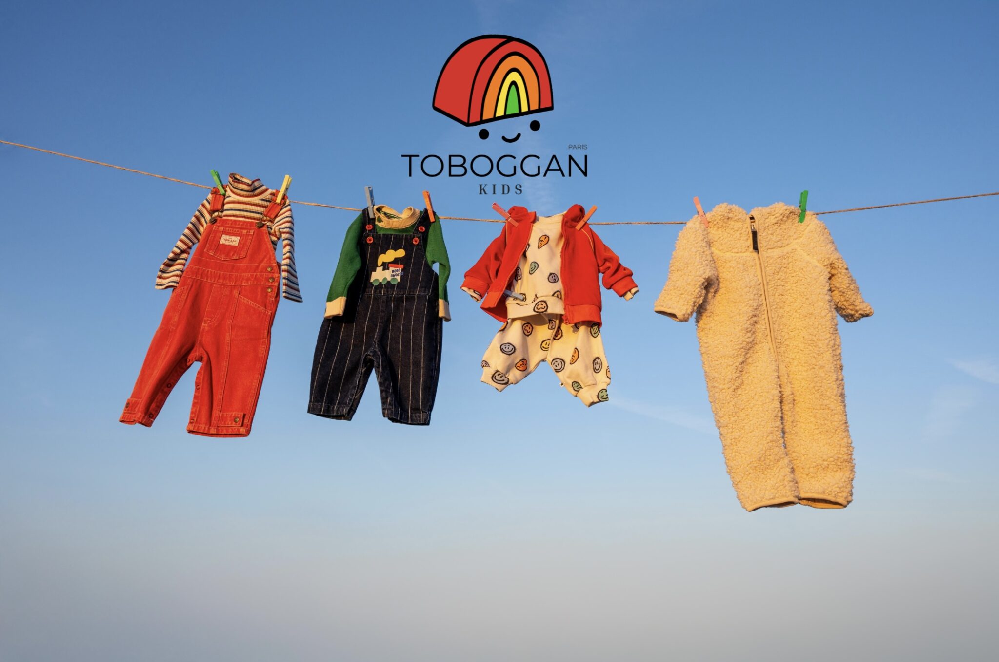 Four colorful kids’ outfits from the online shop Toboggan Kids hanging on a clothesline against a bright blue sky, featured on Scimparello Magazine. Looks include red corduroy overalls with a striped top, dark denim overalls with a green sweater, a playful printed set with a red cardigan, and a cozy cream teddy onesie. A cheerful, outdoor scene full of color and childhood energy.