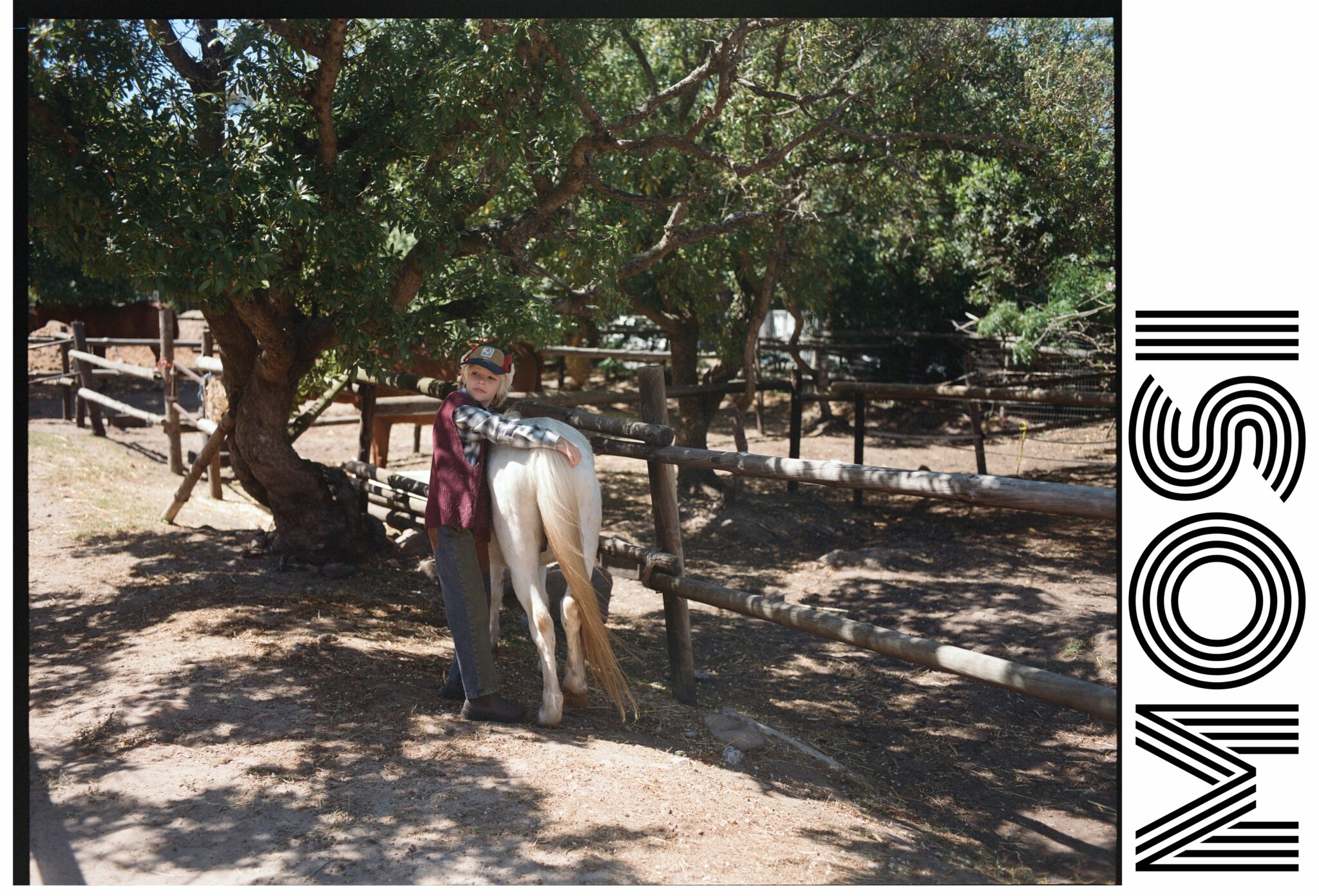 Child hugging a white pony in a sun-dappled paddock, wearing a layered countryside look for the kids fashion editorial Mosi. Photo by Alessandro Iovino, styling by Ulrike Kache, for Scimparello Magazine.