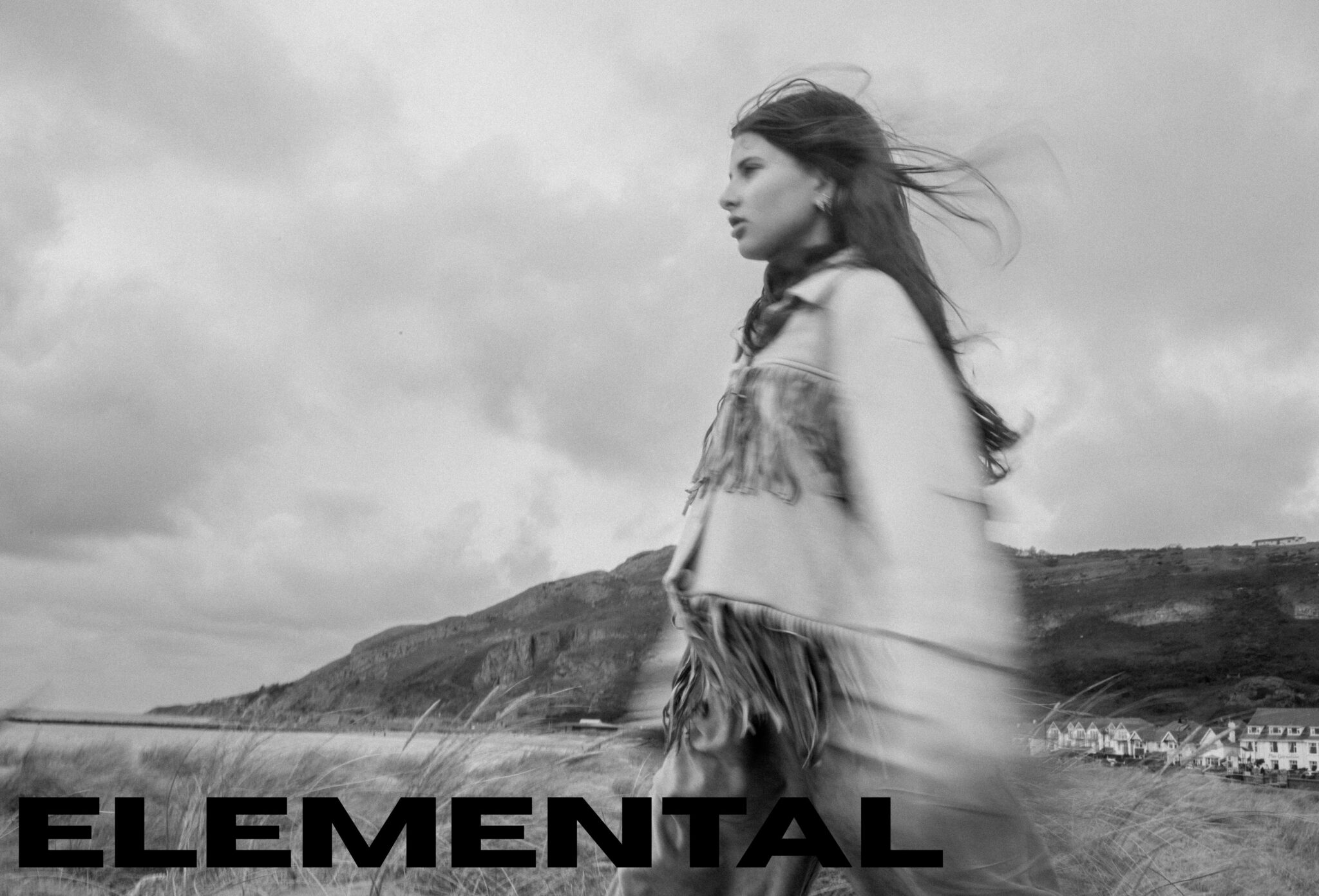 Black and white kids fashion editorial Elemental featuring a girl walking through a windswept coastal landscape, wearing a fringed jacket, photographed by Hannah Mentz for Scimparello Magazine.